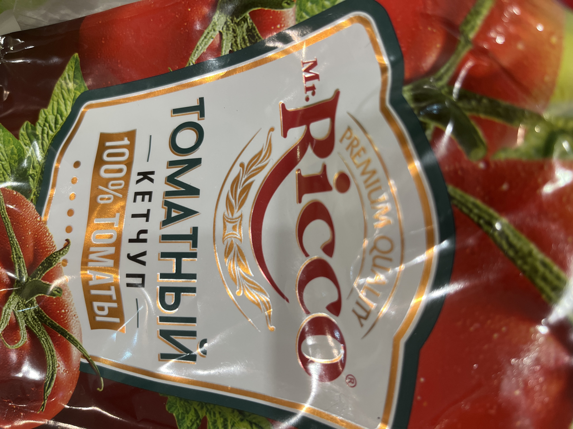 The image displays a close-up, angled view of a flexible food product package for Mr. Ricco Tomato Ketchup, located in Tyumen, Russia. The package label is white, framed by a dark green outer border and a gold inner border. The prominent brand name "Mr. Ricco" is rendered in stylized red capital letters, with a smaller "Mr." preceding "Ricco." Above the brand, a gold decorative crest-like emblem is visible. Below the brand, the phrase "PREMIUM QUALITY" is inscribed in smaller gold capital letters. Russian text on the label reads "ТОМАТНЫЙ КЕТЧУП" (Tomato Ketchup) and "100% ТОМАТЫ!" (100% Tomatoes!), presented in dark green and gold lettering respectively. The background of the package features an out-of-focus illustration of ripe red tomatoes with green stems and leaves. The surface of the packaging is glossy, reflecting ambient light. No individuals or specific actions are depicted in the scene.