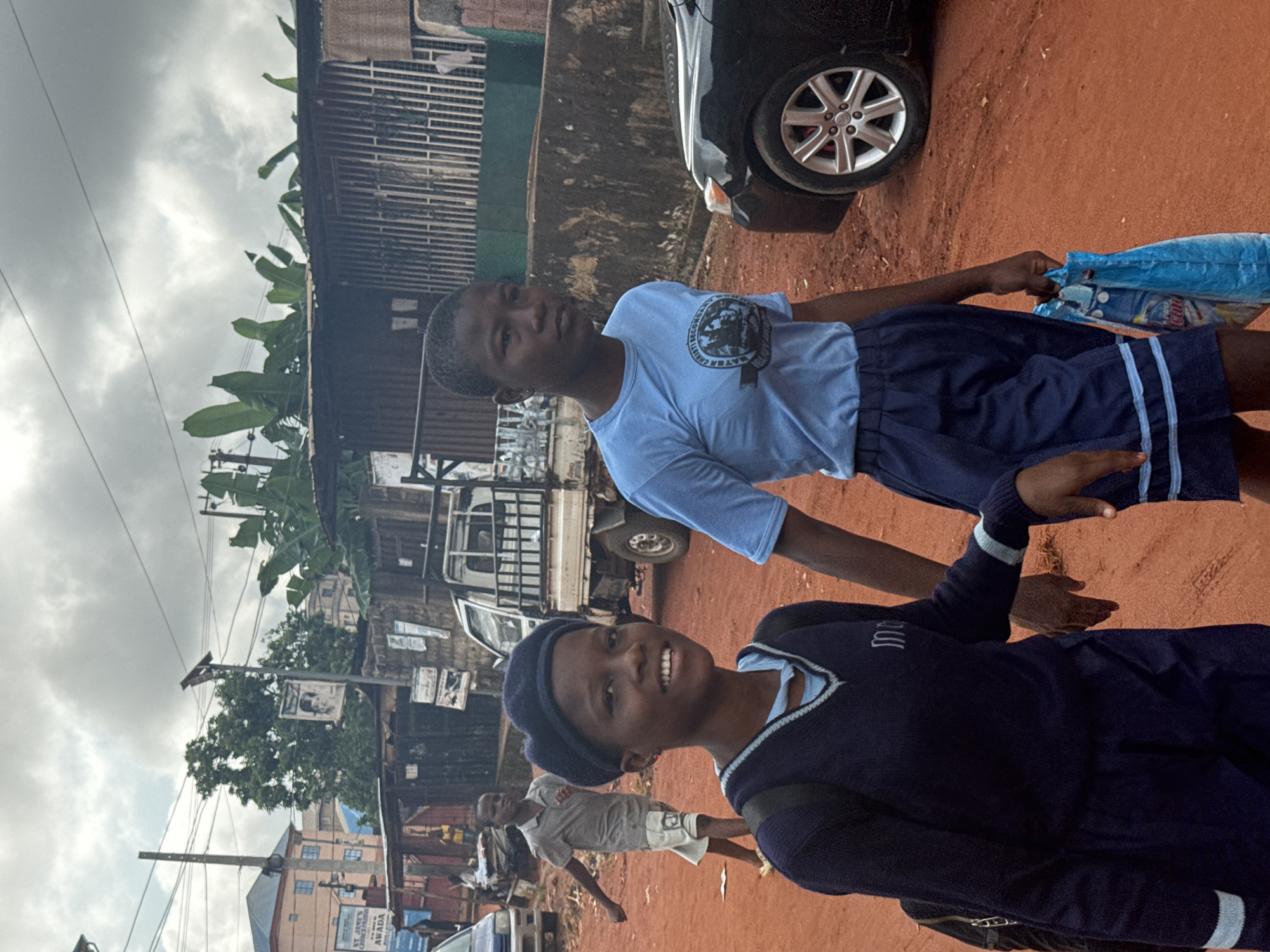 In Onitsha, Nigeria, two young female students walk on a reddish-brown dirt road under a cloudy sky. The student in the foreground, on the left, wears a dark blue v-neck sweater with light blue trim over a light blue top and a dark blue beret. She smiles broadly, holding hands with the student next to her, whose arm is not fully visible. The second student, on the right, wears a light blue t-shirt with a circular emblem and dark blue shorts with light blue stripes. She carries a blue plastic shopping bag in her right hand. In the background, a white truck is parked, and several buildings, including a multi-story structure with blue trim and a corrugated metal structure with bars, line the street. Other pedestrians and a black car are also visible.