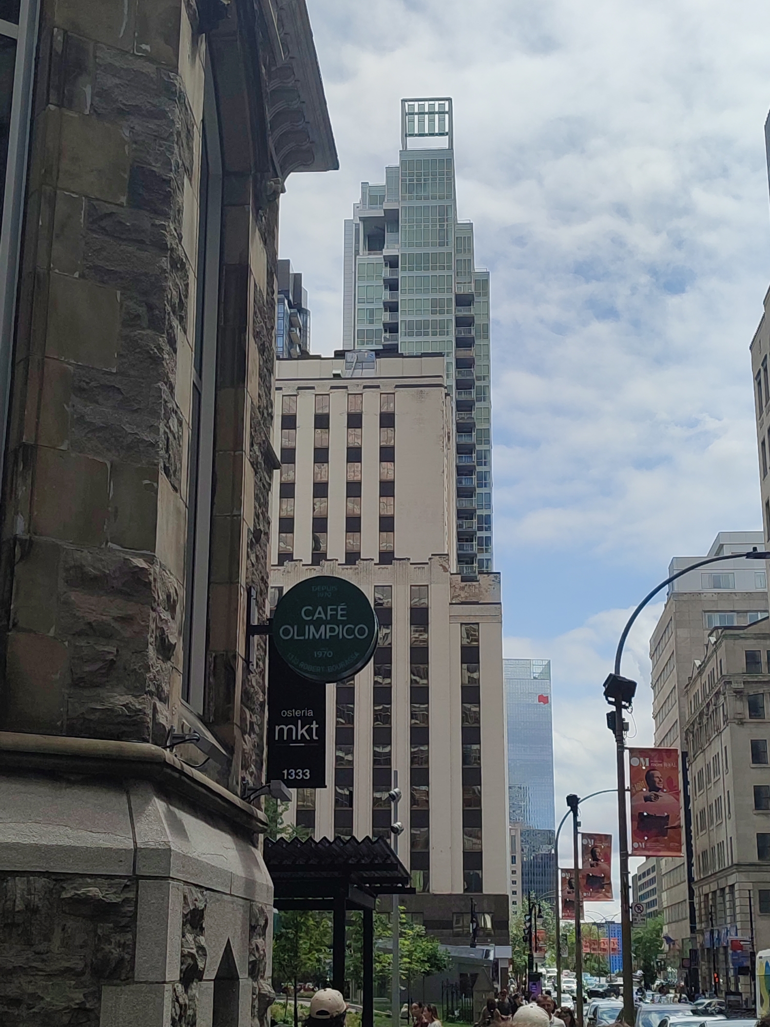 The image captures a street scene in Montreal, Canada, with a focus on the architecture and streetscape. In the foreground, a stone building with ornate architectural details is visible on the left. Adjacent to it, a dark green, circular sign for "CAFÉ OLIMPICO" is mounted, with smaller text below indicating "osteria mkt" and "1333". Behind these elements, a tall, cream-colored building with rows of windows stands, and further in the background, a modern, glass skyscraper dominates the skyline. The sky is partly cloudy. Several streetlights with banners are present, and a busy street with vehicles and pedestrians is visible in the distance. The overall impression is a dynamic urban environment that blends historic and contemporary architecture.