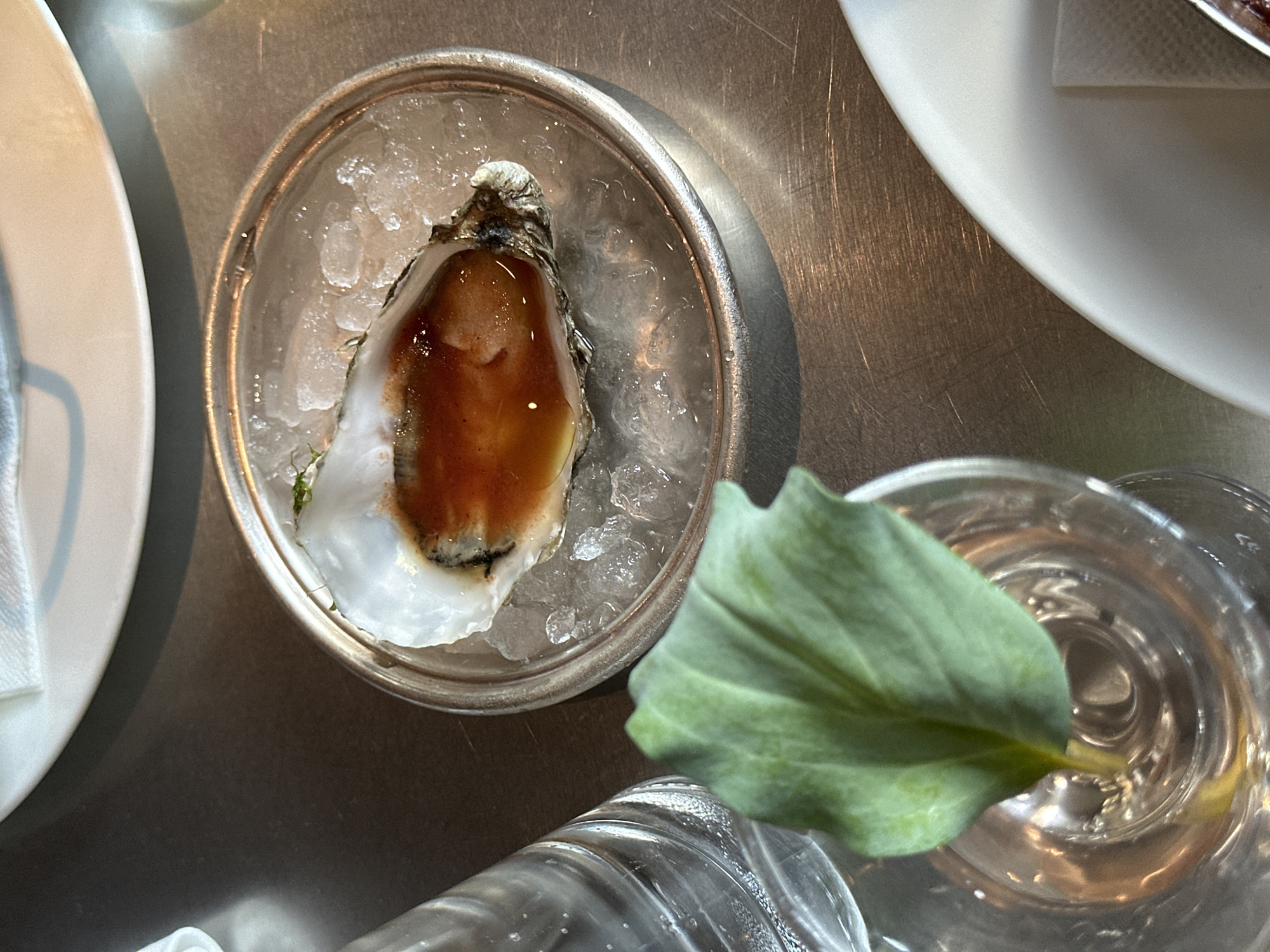 The image shows an oyster served in a silver dish filled with ice, topped with a dark sauce. A leaf partially covers a glass of water. The scene is likely indoors, on a metallic surface, possibly a restaurant table. Two white plates are partially visible at the edges of the image. There are no people visible. The precise location in London is not identifiable from the image.