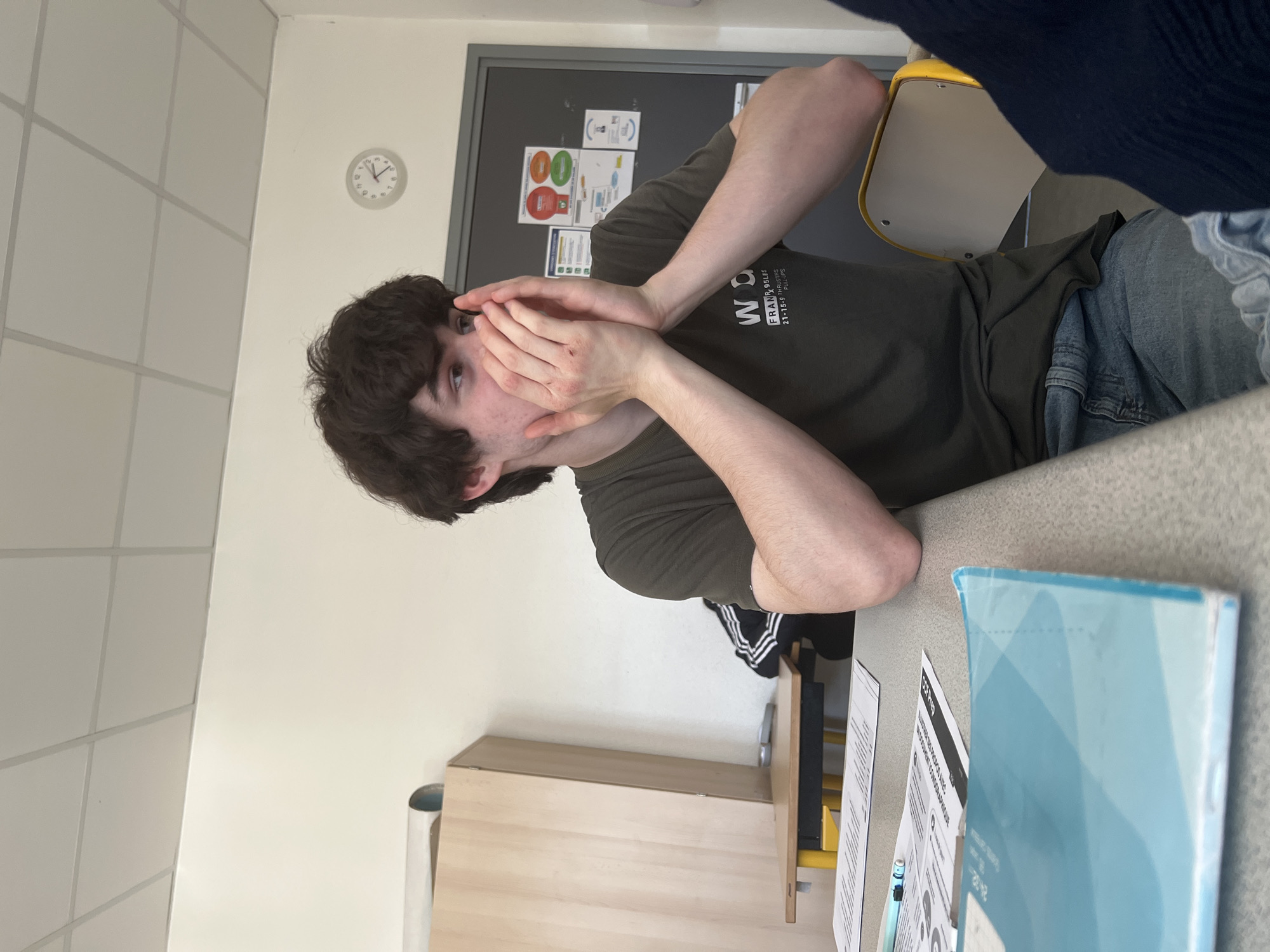 A young man with dark curly hair sits at a desk, his hands near his face. He wears a dark green t-shirt and blue jeans. Behind him is a gray bulletin board with various papers. A clock is visible on the wall. The desk has papers and a book on it. The environment appears to be a classroom. It is not possible to determine the precise location in Schiltigheim from the image.