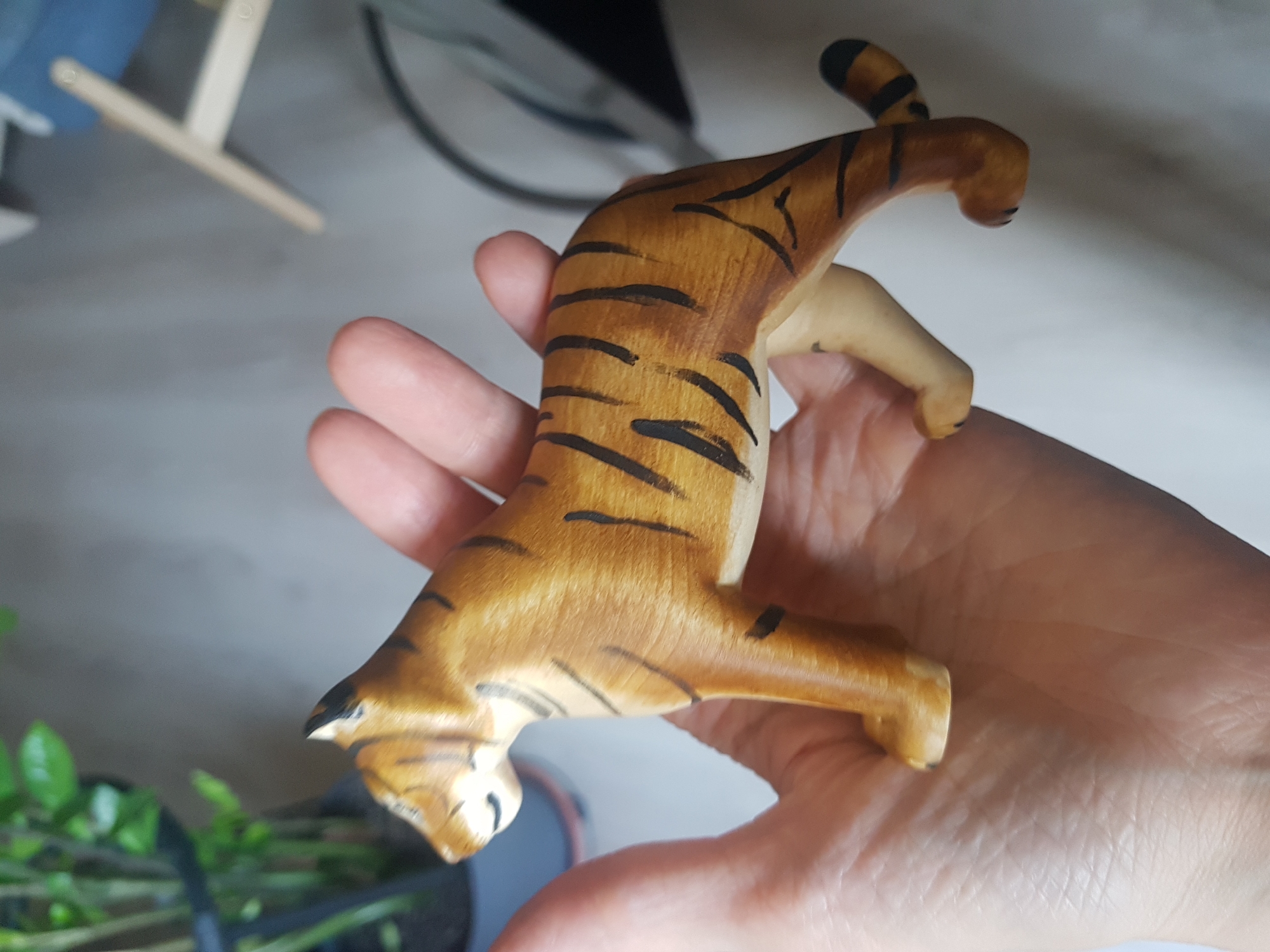 A hand is holding a wooden tiger figurine. The tiger is painted with orange and brown stripes. The background is blurry, but includes a plant, a table, and a chair. The location is not discernible from the image, and it is not possible to identify the location as Saint Petersburg, Russia.