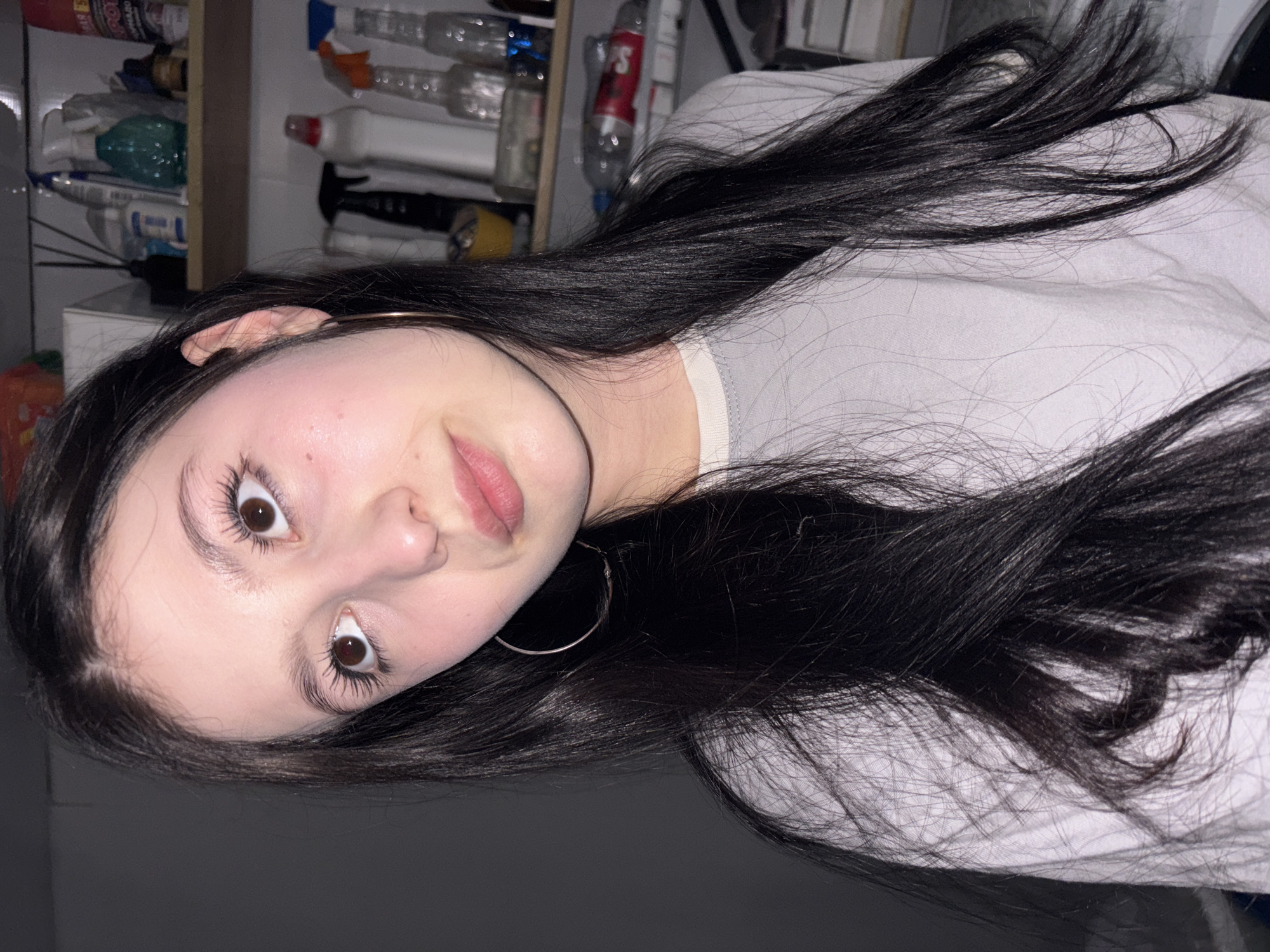 A woman with dark hair and brown eyes is pictured from a close-up perspective. She is wearing a light gray shirt and a silver hoop earring. The background is indistinct, but a shelf with various bottles and containers is visible. The location is not specified.