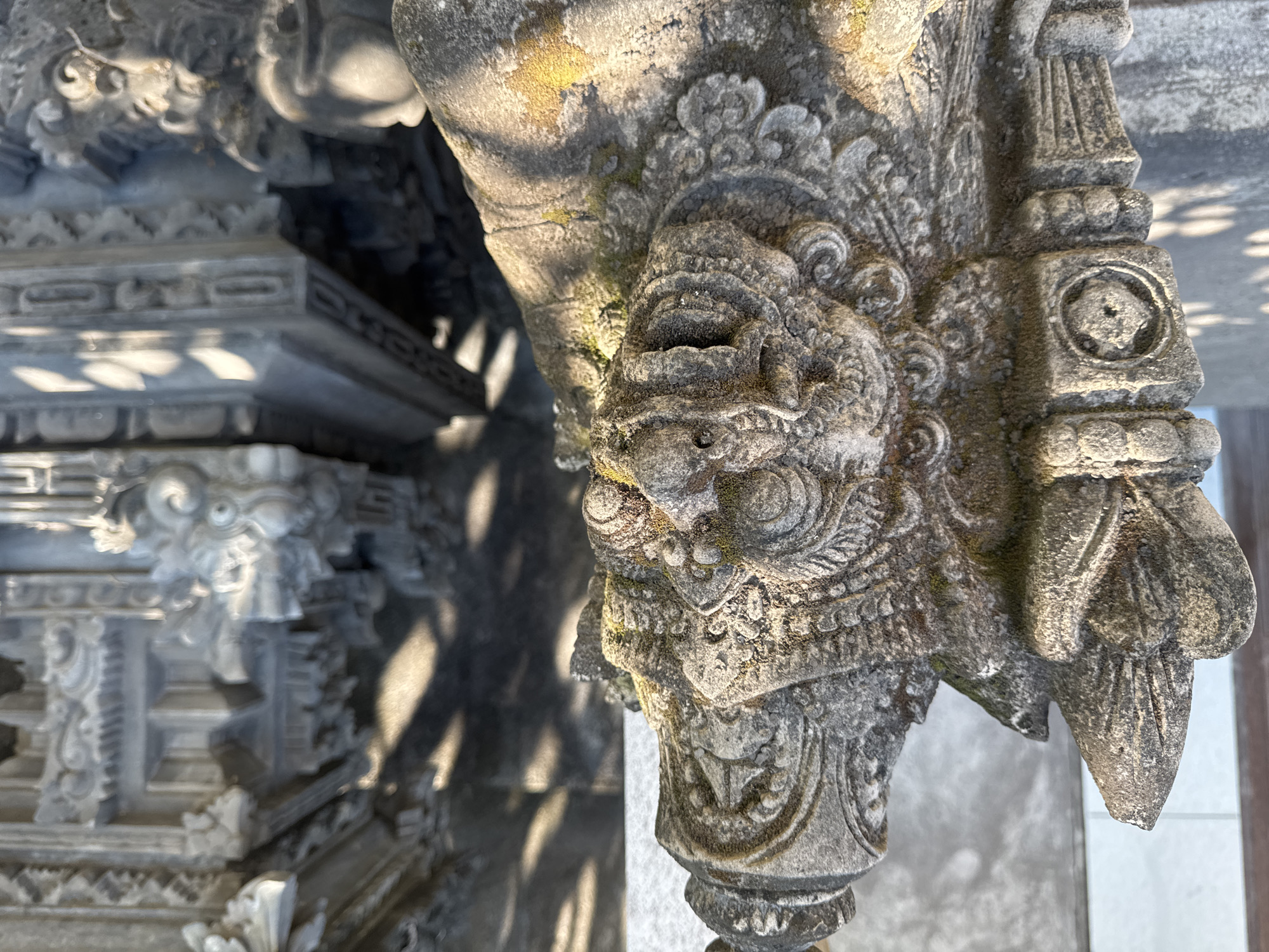 The image depicts a close-up of a weathered, intricately carved stone structure, likely part of a temple or ancient building. The primary focus is on a carved lion's head with detailed ornamentation. The stone is gray and shows signs of age, with moss and discoloration in some areas. The background shows other parts of the carved structure in soft focus, with geometric patterns and details. The light casts shadows, highlighting the texture of the stone. The location is Kubu, Indonesia.