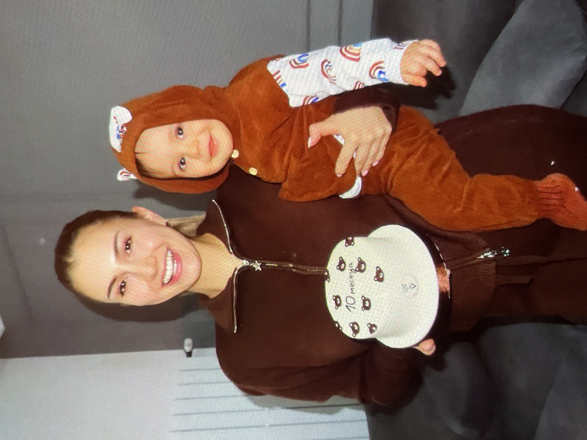 A woman is holding a baby in a brown bear outfit. She is also holding a white plate decorated with bear images and the number "10" in what appears to be Russian text, likely indicating 10 months. The woman is smiling and wearing a brown jacket or sweatshirt. The background includes a gray wall and a dark-colored couch. Based on the provided information about location, this scene is in Harmanli, Bulgaria.