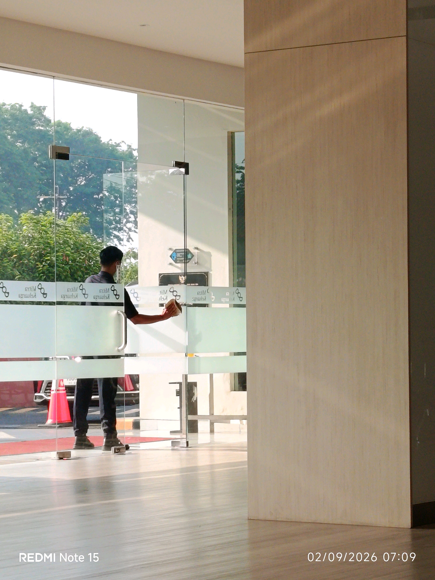 In Waru, Indonesia, a man wearing black pants and a black short-sleeved shirt is cleaning a glass door with a light-colored rag. He is positioned in the doorway, facing away from the camera, and his left arm is extended as he wipes the glass. The glass door has horizontal frosted bands with a repeating logo or pattern. Outside the glass, green trees are visible. The floor inside appears to be polished wood or tile, reflecting the light. A large, light-colored pillar or wall is to the right of the frame. The scene is illuminated by natural light, creating shadows on the floor. The timestamp "02/09/2026 07:09" and the text "REDMI Note 15" are visible in the lower portion of the image.