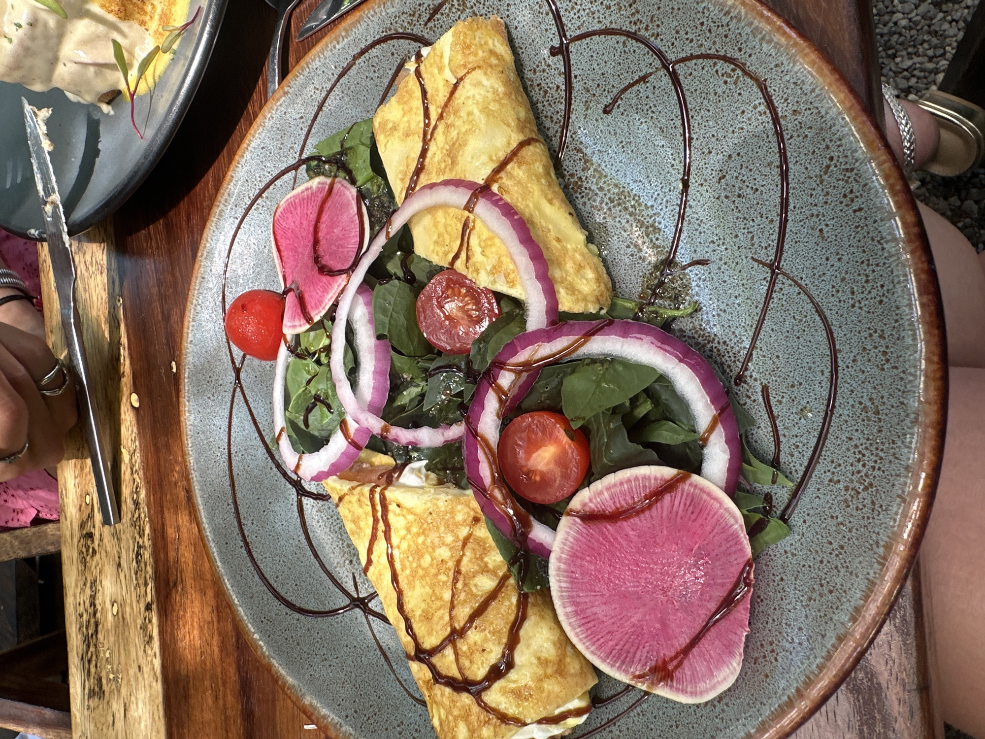 A top-down shot captures a plate of food on a wooden surface. The plate, of a rough ceramic with brown lines forming a circular pattern, holds two folded omelets drizzled with a dark sauce. Slices of pink radish and red onion rings are scattered around the omelets, along with cherry tomatoes and green leaves. A knife held in a person's hand is visible to the left of the plate. A woman's legs are visible in the top right. The image was likely taken at a restaurant in Acapulco de Juárez, Mexico.