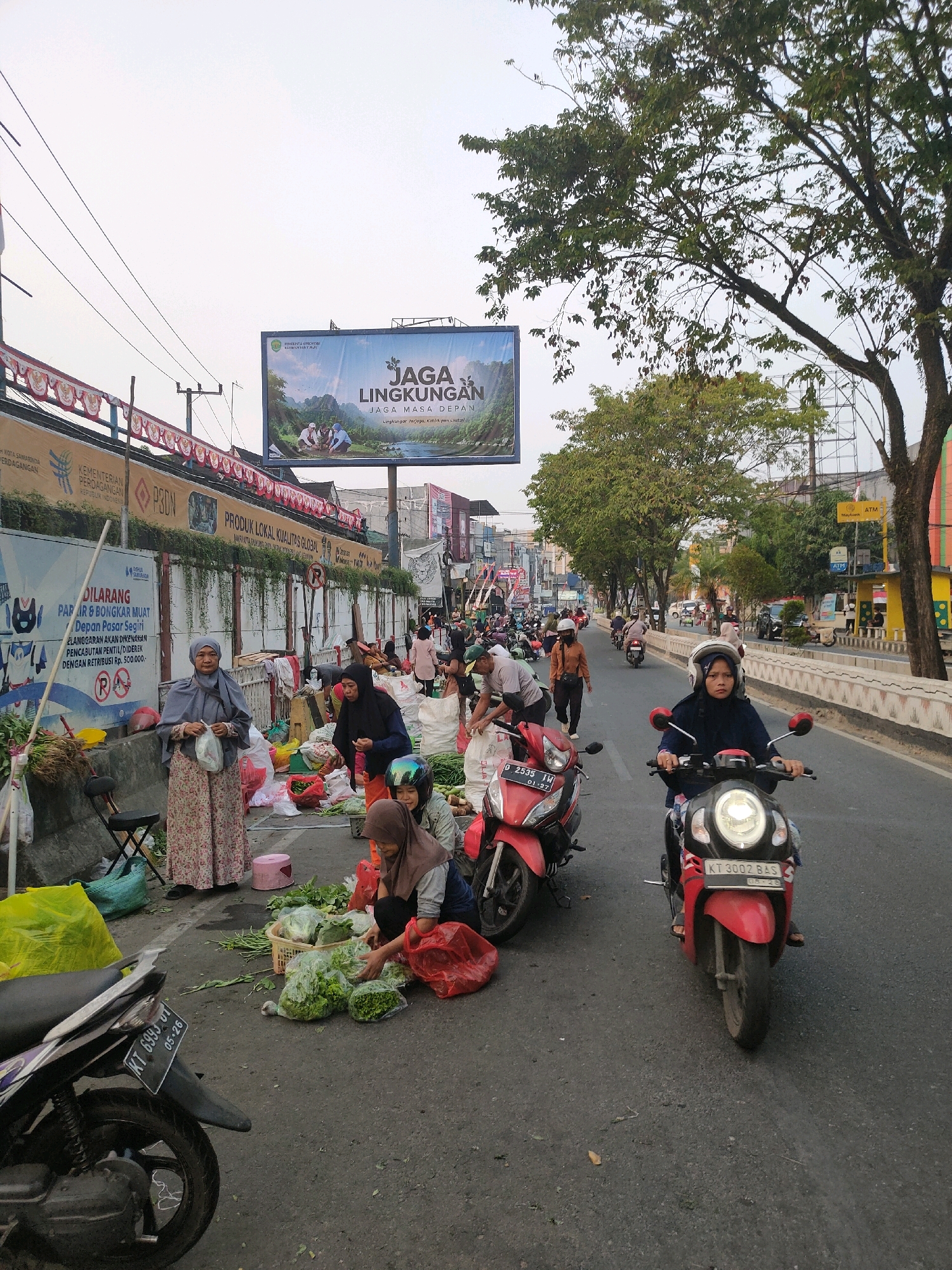 The image depicts a bustling street scene in the city of Samarinda, Indonesia, likely a marketplace. In the foreground, a woman on a red scooter is riding down the street, wearing a blue jacket and a helmet. To her left, a red scooter is parked. The street is lined with vendors selling produce, with several women in headscarves visible among the stalls. Bags of vegetables are piled on the ground, and some are displayed in baskets. In the background, there are buildings and trees, and a large billboard with Indonesian text about environmental preservation. The sky is overcast, suggesting it might be early morning or late afternoon. The overall atmosphere is one of everyday commerce and activity in a Southeast Asian urban setting.