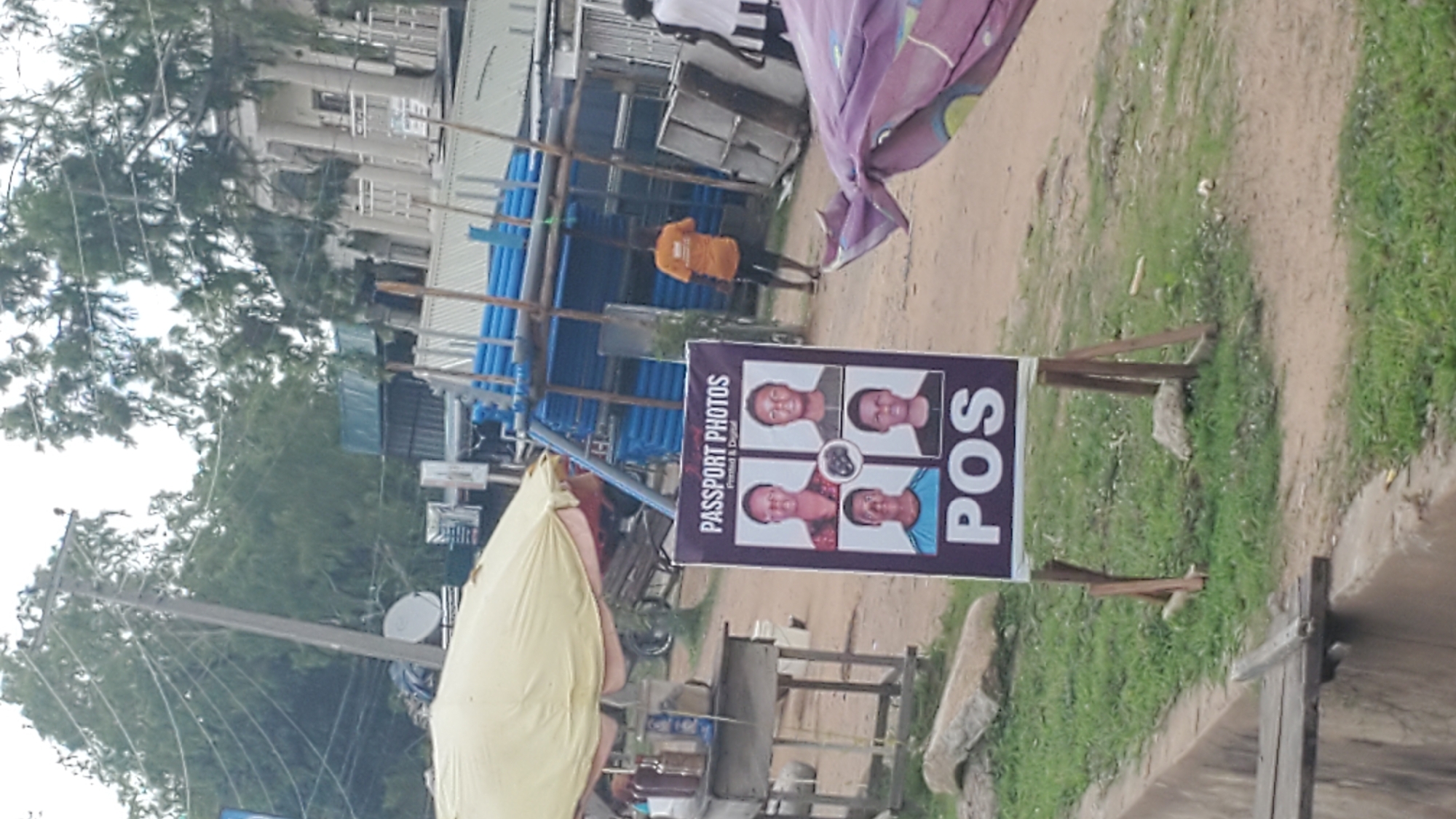 A passport photo booth stand is set up outdoors in Jalingo, Nigeria. The stand is a purple sign with "PASSPORT PHOTOS" printed at the top, followed by four passport-sized photos of different individuals and "POS" at the bottom. To the left of the stand, there is a table with bottles and a yellow umbrella providing shade. In the background, there are stacks of blue pipes, some structures with metal roofs, and a white building with columns. A man in an orange shirt is visible walking in the background. The environment appears to be a dusty, open area with some grass.