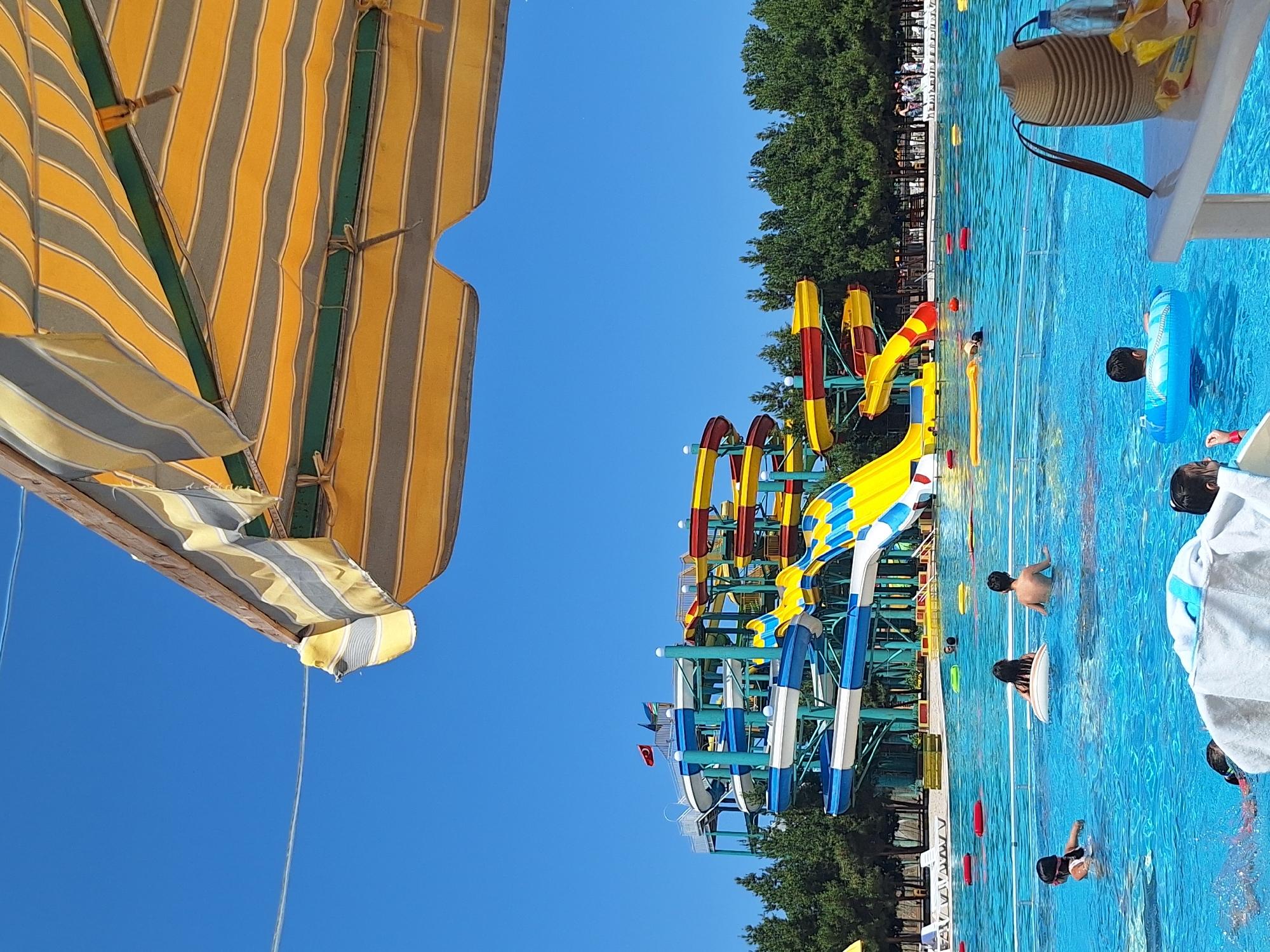 The image depicts a sunny day at a water park in Khudat, Azerbaijan. In the foreground, a portion of a striped yellow and grey awning is visible, casting a shadow over the scene. The background is dominated by a large, multi-colored water slide complex with blue, white, yellow, and red sections, towering above a clear blue swimming pool. Several children are visible in the pool, some swimming and others floating on inflatable rings. Red and yellow inflatable buoys are scattered across the water. A table with a straw bag, a water bottle, and some snacks is in the lower right corner. Trees line the perimeter of the water park, and a clear blue sky completes the scene.