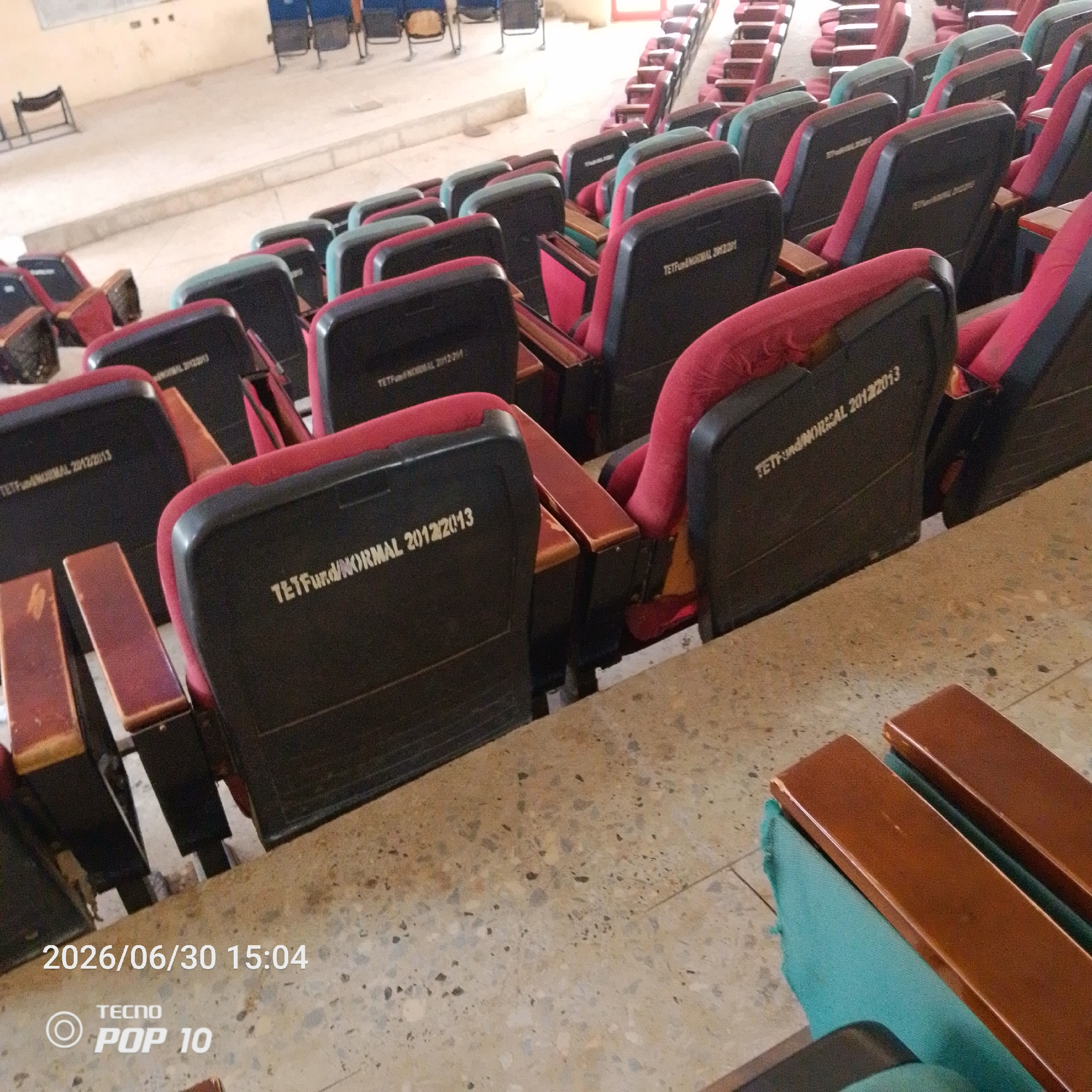 This media file depicts an interior scene of an empty lecture hall or auditorium in Jalingo, Nigeria. The primary subjects are rows of empty seats, some with red and black upholstery, and others with teal upholstery. The seats are arranged on tiered platforms, indicating a large gathering space. The floor is a speckled terrazzo material.
Visible text on the seats indicates "TETFund & NORMAL 2012/2013" and "TETHONG/NORCAL 2012/2013", suggesting these seats were funded or are part of a program associated with these entities and a specific academic year. A watermark in the bottom left corner displays "2026/06/30 15:04" and "TECNO POP 10", which is likely the date and time the photo was taken, and the device used.
The scene is brightly lit, suggesting it is daytime. There are no people visible, and the overall atmosphere is still and quiet, implying the space is currently unoccupied, perhaps between events or classes. The setting appears functional and educational, consistent with a university or college environment. The image focuses on the details of the seating and the floor, with a slightly blurred background showing more rows of seats and a distant wall.