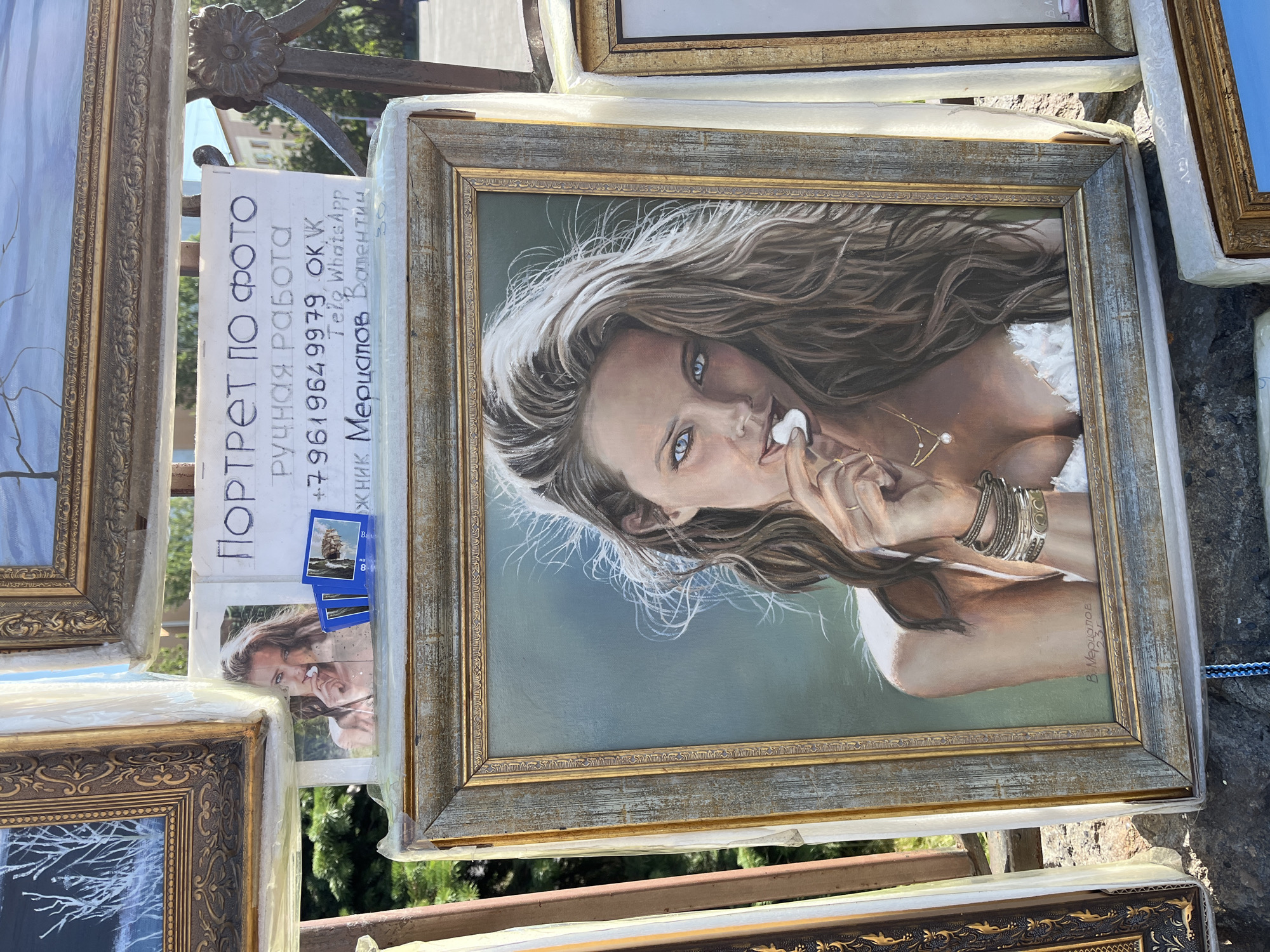 The image shows a street art display in Petropavlovsk-Kamchatsky, Russia. In the foreground, a framed oil painting of a woman with blue eyes and long brown hair is displayed. She is wearing a white top and has a small white object in her mouth. The painting is signed "В. Мерцалов" and what appears to be "237".
Behind the painting, a handwritten sign advertises "PORTRAIT FROM PHOTO" and "HANDMADE". It also provides a phone number, "+79619649979", and lists "OK, VK, Tele, WhatsApp". The name "Мерцапов Валентин" is also written on the sign. A small photograph of a woman is attached to the sign.
Several other framed artworks are visible in the background, including a painting of trees in a snowy landscape. The display is set up outdoors, with what appears to be a stone wall in the lower part of the image and greenery in the background. The overall impression is that of an artist displaying their work for sale on the street.