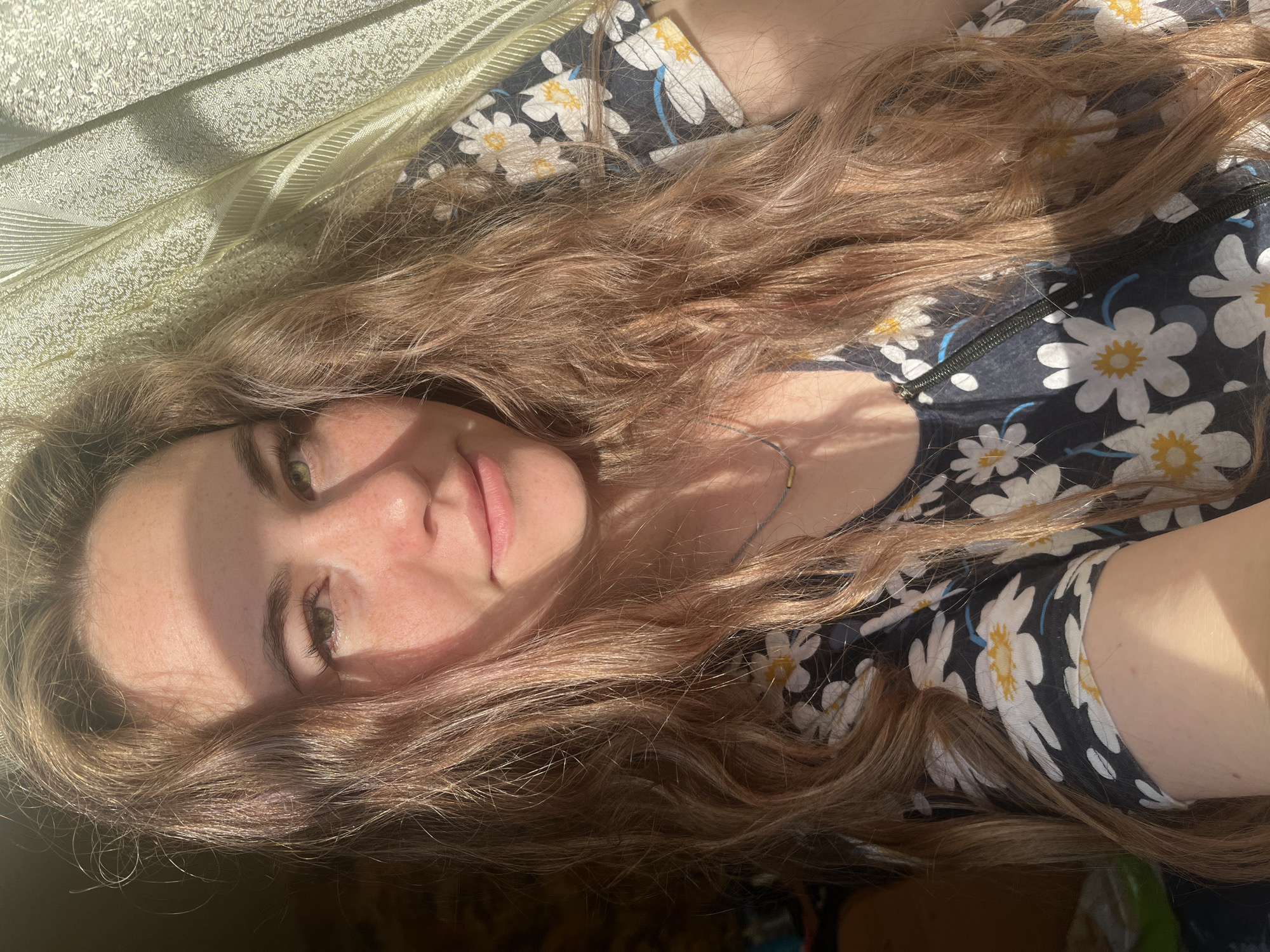 A young woman with long, wavy brown hair and fair skin is lying down, facing the camera. Her head is tilted to the side. Her face is partially illuminated by sunlight, creating strong shadows on one side. She has green eyes, visible freckles, and pink lips. She is wearing a dark blue floral print top with white daisies with yellow centers. Part of a light-colored, textured surface (possibly a blanket or sheet) is visible near the top left of the frame. The location in Сосново, Russia is not identifiable from the given visual information.