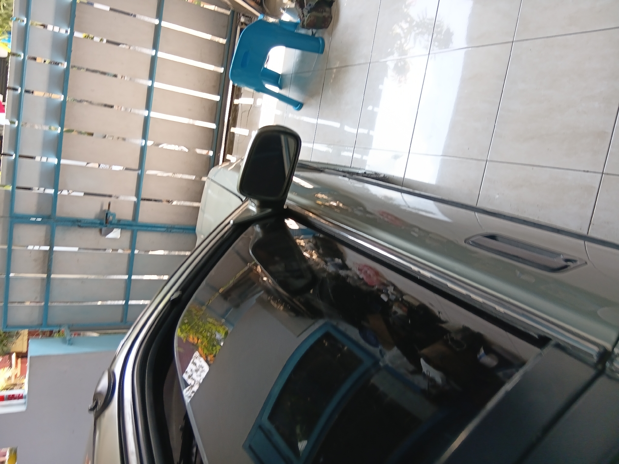 The image, taken in Tanggul Wetan, Indonesia, shows the side of a light green car parked on a tiled floor, possibly a garage or covered outdoor area. The car's window and side mirror are visible, reflecting the surroundings. Behind the car is a light blue gate or fence with vertical white slats. To the right of the car, a bright blue plastic stool is placed on the tiled floor, and a dark bag or object is situated near the fence. The lighting suggests it is daytime, with sunlight casting reflections on the car and floor.