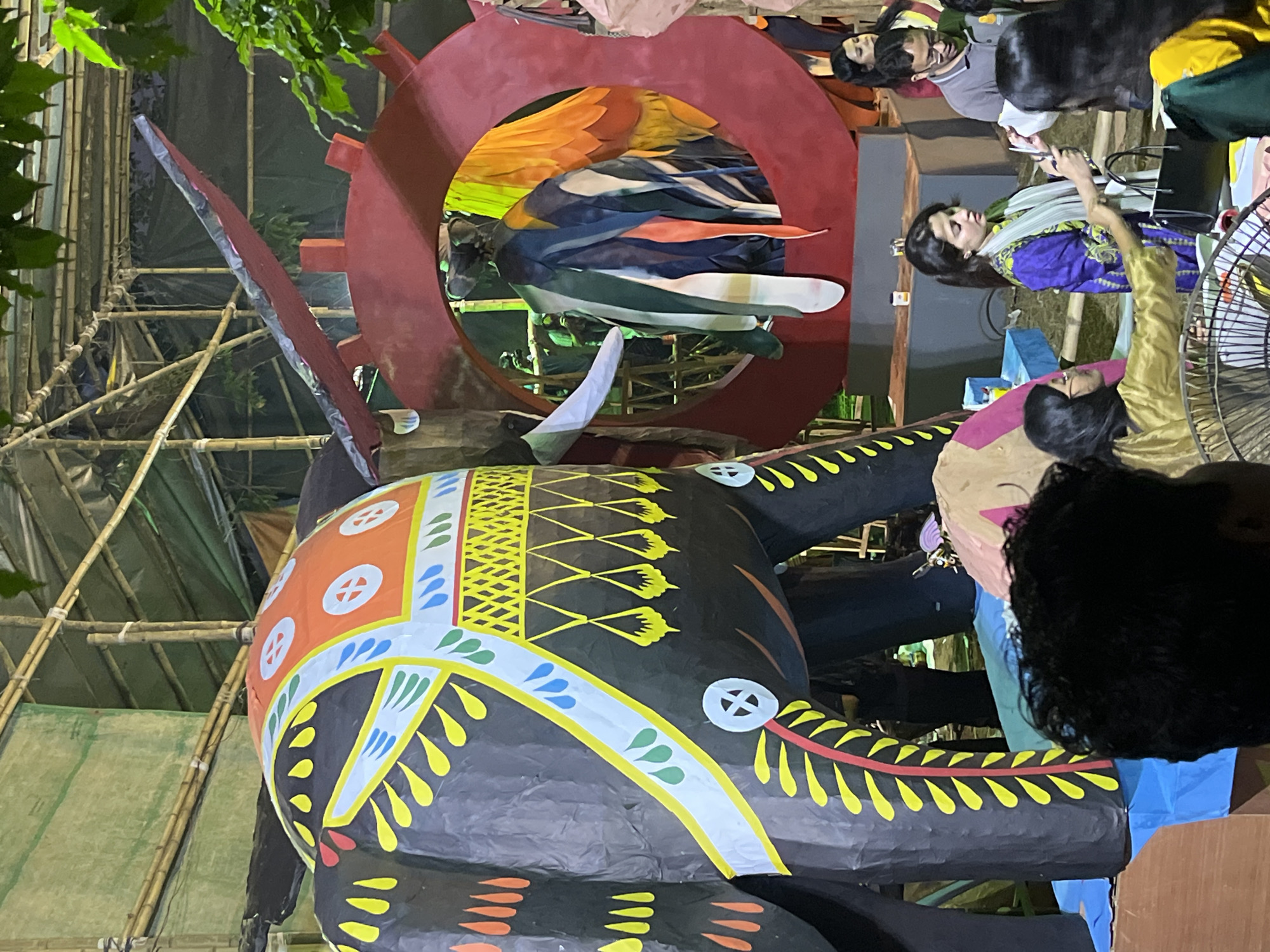 The image captures a street scene in Dhaka, Bangladesh, likely during a festival or event. At the center, a large, brightly painted elephant sculpture is prominent, featuring black, orange, yellow, and white geometric patterns. The sculpture is positioned in front of a red circular structure containing colorful fabrics or materials.
Several people are visible in the frame. A woman dressed in a purple and gold outfit with a white shawl is looking downward at something, possibly a device or another person. Another person in a gold-colored garment is partially visible next to her. Other individuals are observed standing in the background.
A bamboo scaffold structure is present behind the elephant sculpture, and some green foliage or trees are seen overhead. The lighting suggests it is either dusk or night, with artificial lighting illuminating the scene. There is a sense of community and festivity in the setting.