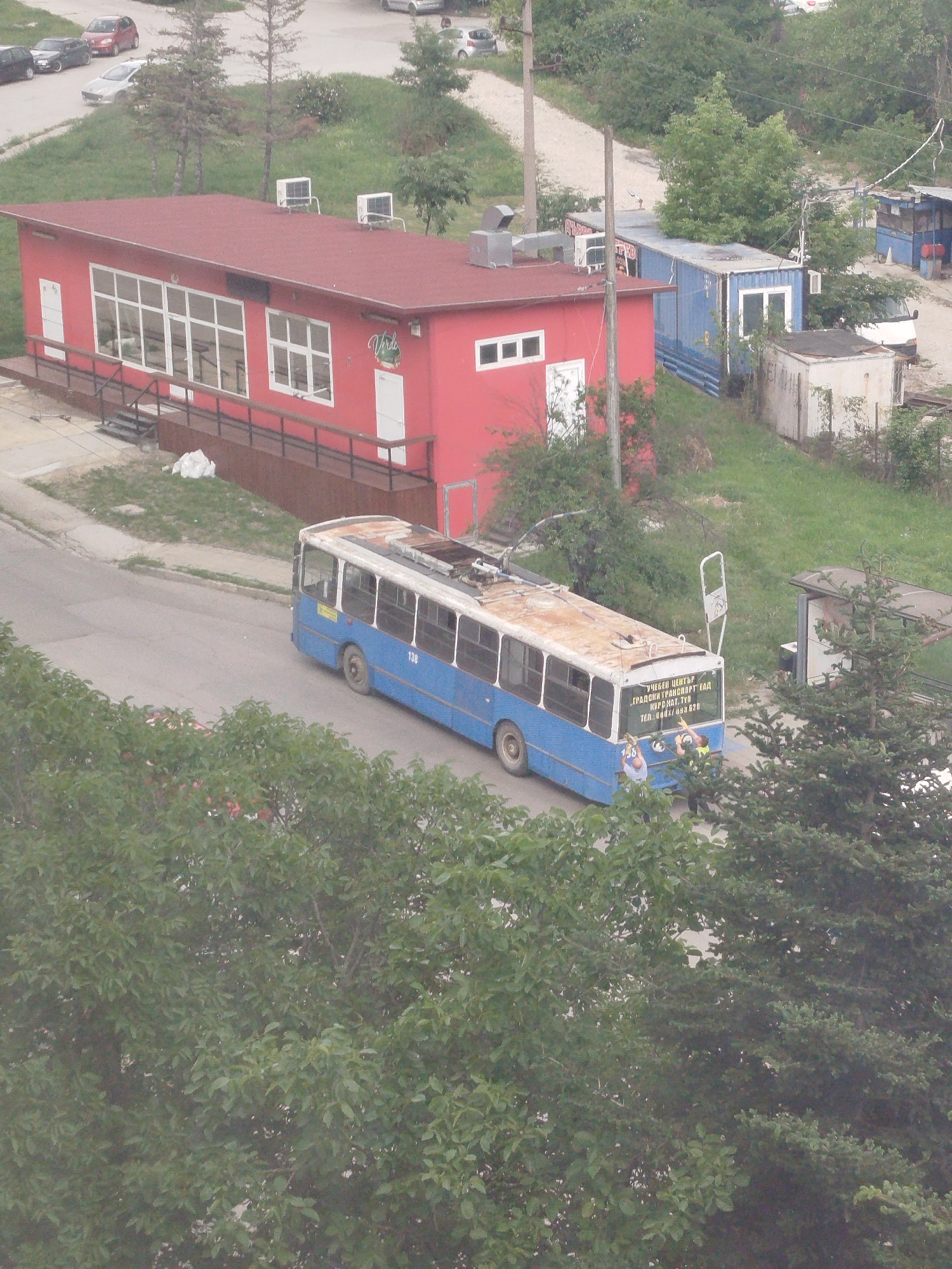 The image depicts a street scene in Varna, Bulgaria. A blue and white trolleybus, labeled "138", is positioned on the asphalt road. The trolleybus has a text on its back which reads "Учебен център 'Градски транспорт' ЕАД, Kurs hat, Tyo tel. 0002/083 620". Three people are visible near the rear of the bus.
In the background, there's a vibrant red, single-story building, likely a restaurant called "Verde". Additional buildings, including a blue container-like structure, are also visible. The environment is somewhat overgrown, with greenery covering a large portion of the scene.