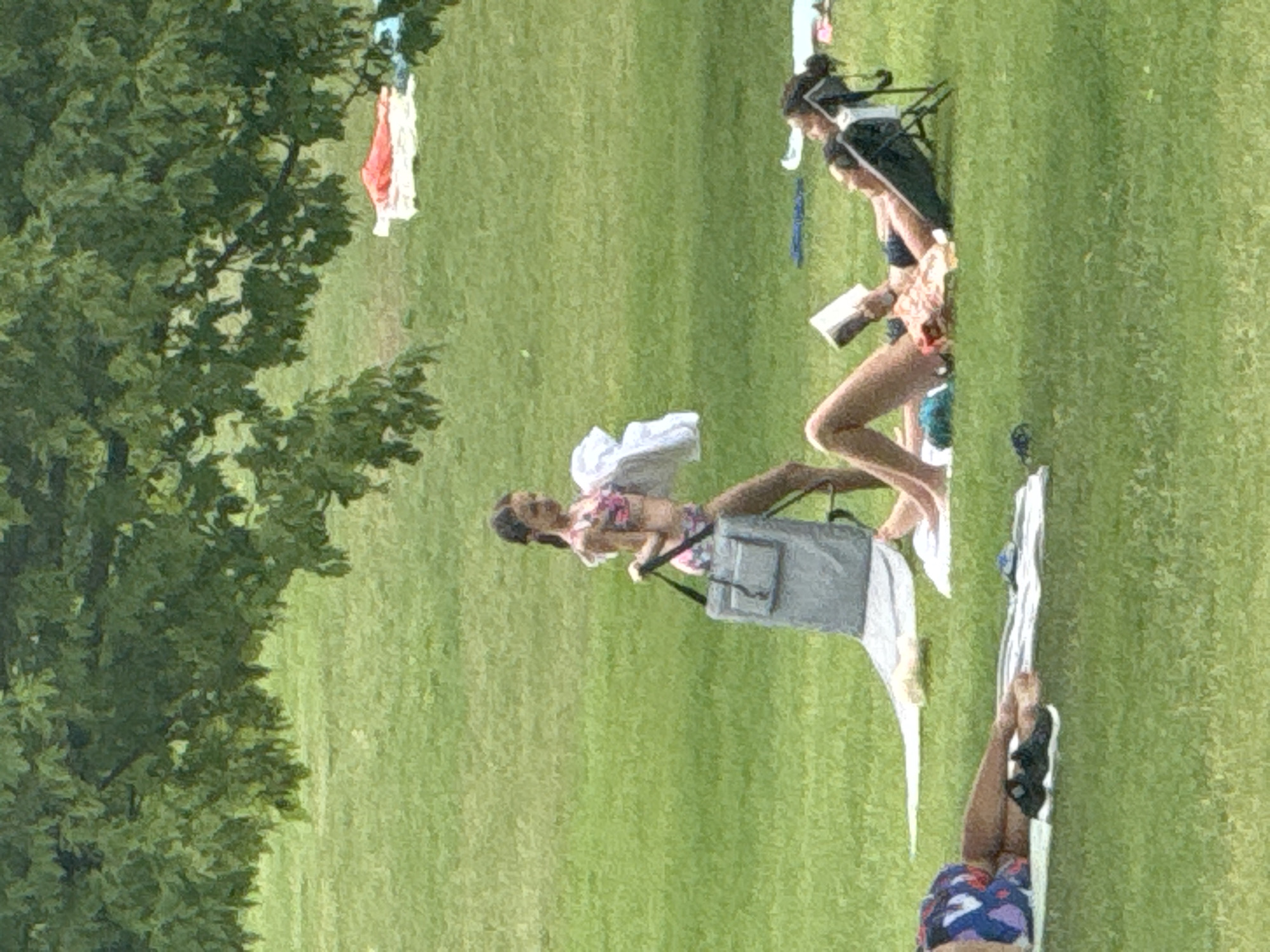 This media file depicts a scene on a grassy field, possibly a park or recreational area. The primary subjects are several individuals, likely women, engaged in leisurely activities. One person is lying on a beach chair, appearing to read a book. Another individual is positioned further away, also seemingly relaxing. In the background, some clothing items, including what appears to be a red and white piece of fabric, are hanging from tree branches, suggesting a casual outdoor setting. The overall atmosphere seems relaxed and summery, with the bright green grass and the relaxed postures of the individuals indicating a pleasant day. The lighting suggests it could be daytime, possibly late morning or afternoon. The presence of beach chairs and what looks like swimwear hints at a possible location near a body of water or a place where people are enjoying warm weather activities. There is no visible text in the image.