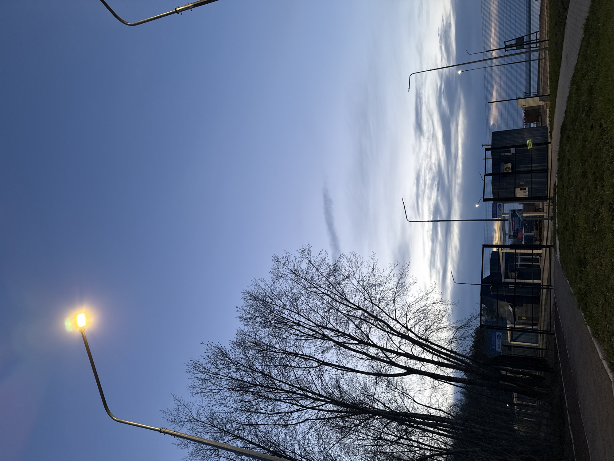 The image shows an outdoor scene in Chaikovsky, Russia. A street lamp is illuminated in the lower-left corner of the frame, contrasted against a clear blue sky. A silhouette of a bare tree occupies the lower portion of the scene. To the right, along a sidewalk, there are buildings and a chain-link fence. The sky transitions into a cloudy appearance towards the horizon, suggesting either sunrise or sunset.