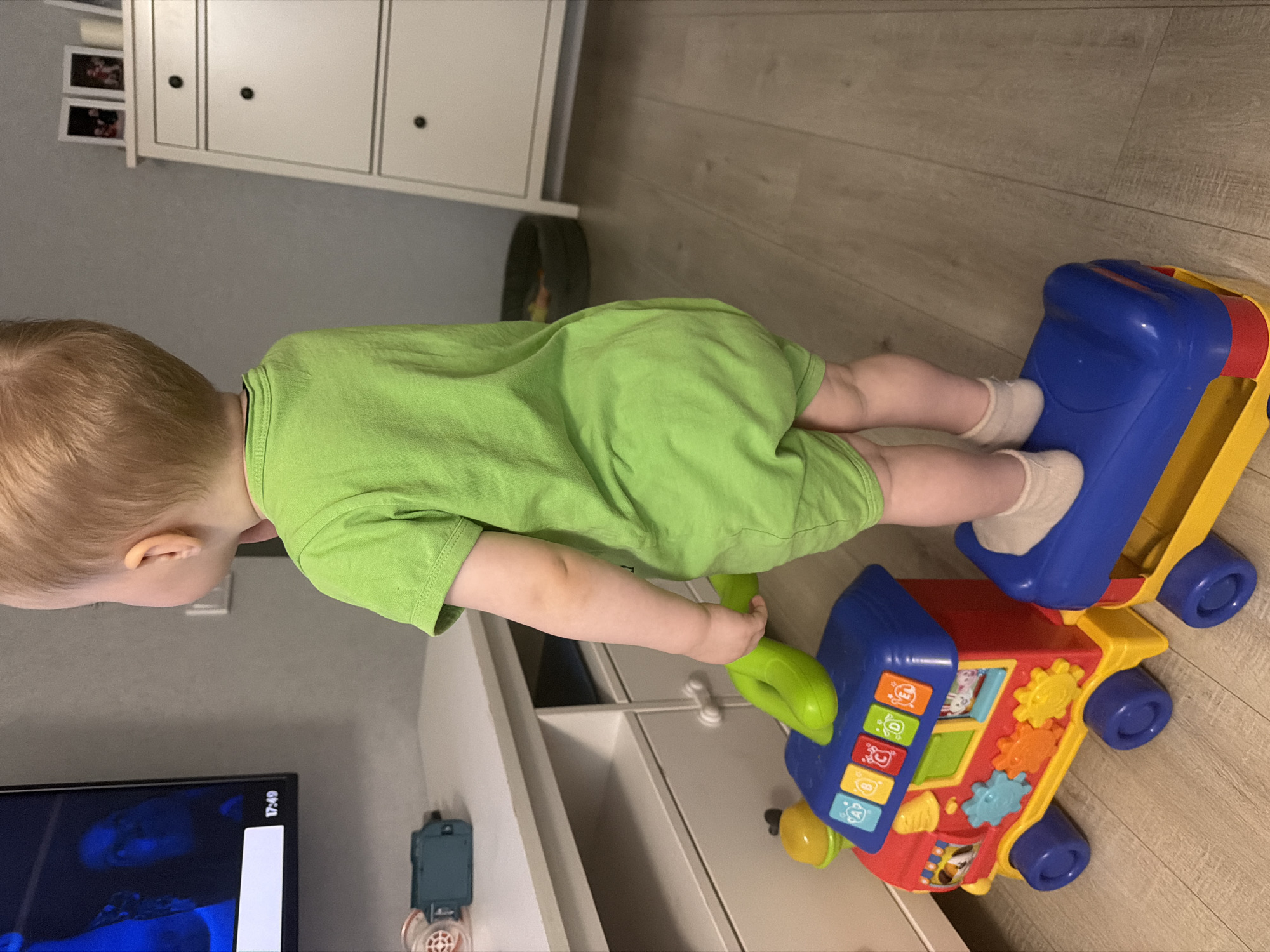 An indoor scene in Votkinsk, Russia, shows a young child, viewed from behind, standing upright on the blue and yellow rear car of a multi-part toy train. The child has light-colored hair, wears a lime green short-sleeved romper, and light-colored socks, and is holding a bright green handle. The child is oriented towards a flat-screen television displaying a dark image with blue hues and a digital time stamp of "17:49" in the lower right corner. The main locomotive of the toy train, positioned on the light-colored wooden floor, is red, blue, and yellow, featuring colorful buttons, gears, and wheels. Beneath the television is a white media console with drawers and open compartments, upon which a green rectangular object and a white and red circular item are placed. To the right of the scene, a tall white chest of drawers with black knobs holds two framed photographs on its top surface. The room features light gray walls, and a dark circular object is partially visible in the background between the console and the chest of drawers.