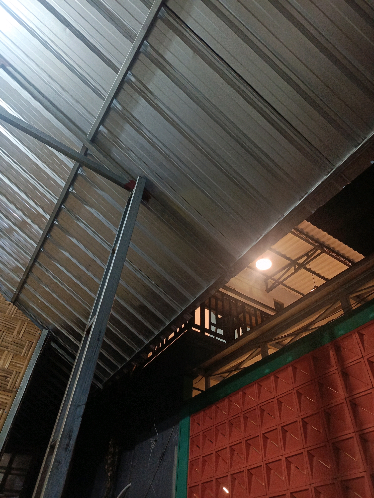 The image shows an exterior view of a building structure with a corrugated metal roof supported by metal beams. The roof angles upward and to the left. Below the metal roof, there are sections of bamboo matting on the left side and a red brick wall with a grid pattern on the right side. A bright lightbulb illuminates a space beneath an overhang, revealing wooden beams and a corrugated ceiling. The environment appears to be outdoors, possibly a covered walkway or a section of an outdoor market or dining area. The lighting suggests it may be evening or a dimly lit interior space.