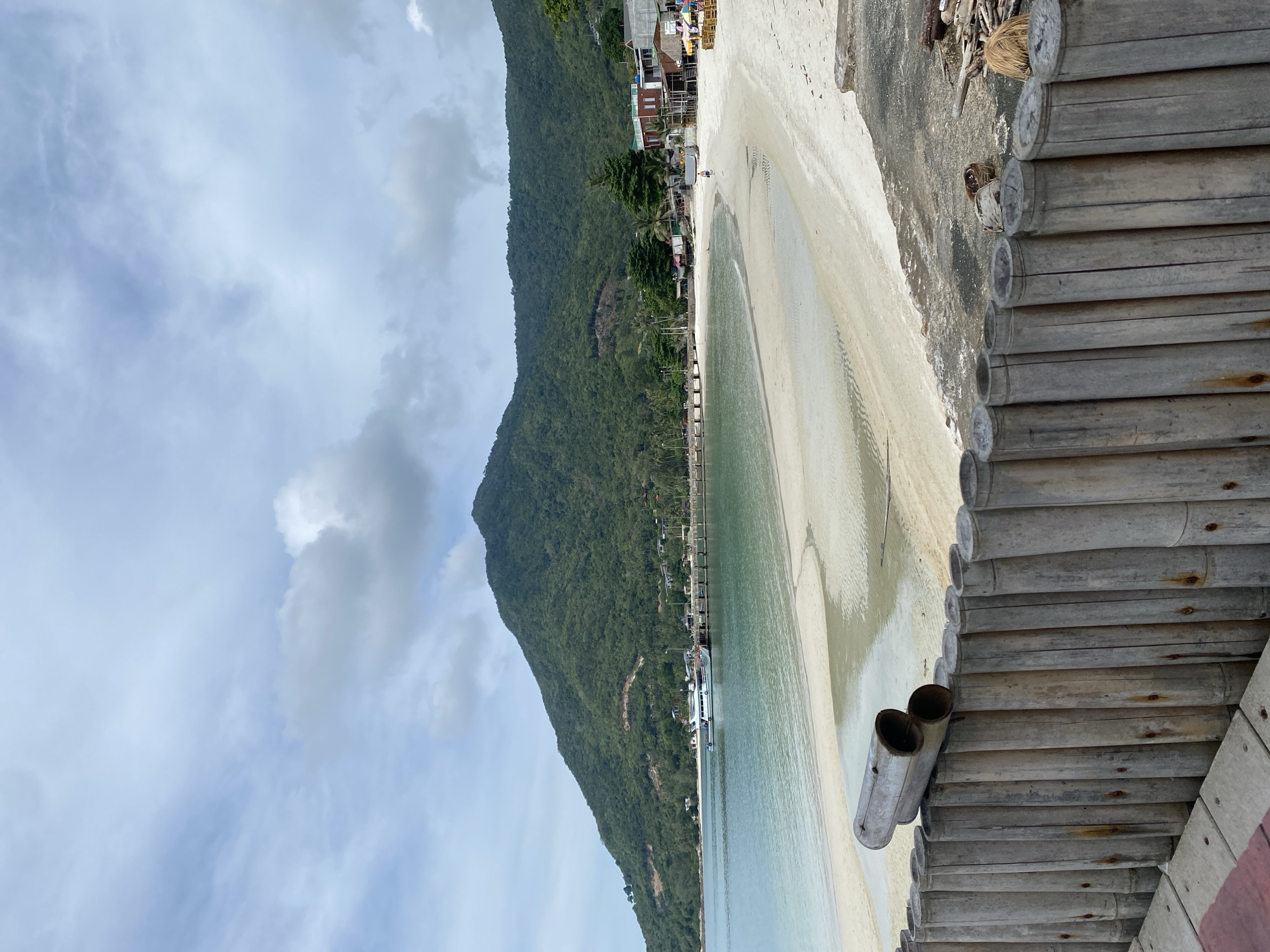The image depicts a beach scene in Baan Chaloklum, Thailand. A lush, green mountain rises along the left side of the frame. The beach itself consists of white sand and shallow, clear water. A pier extends into the water, with several boats docked alongside. The foreground is dominated by a bamboo structure, possibly a walkway or part of a building. The sky is partly cloudy. There are buildings and a few figures visible along the beach near the base of the mountain.