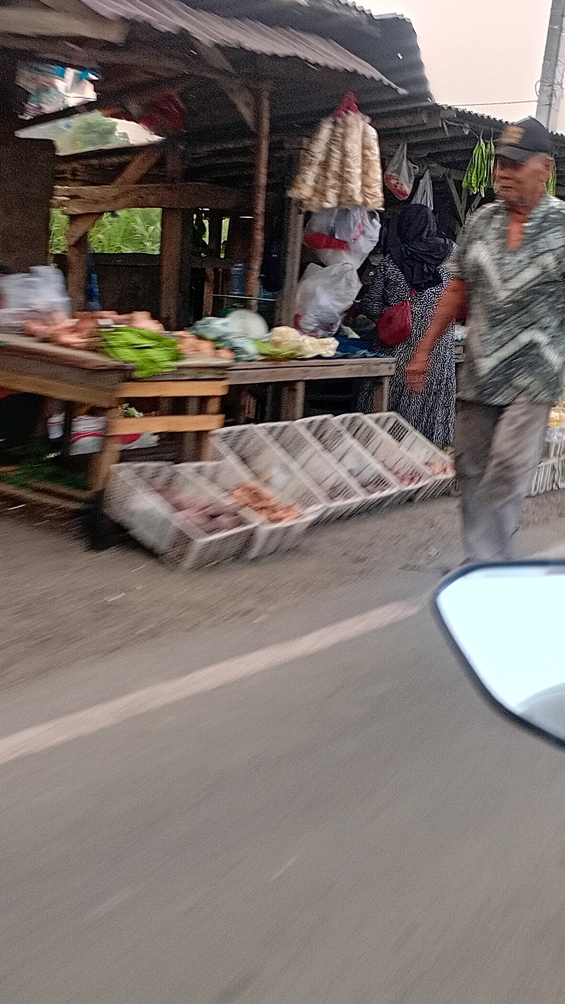 In the city of Jambi, Indonesia, an outdoor market stall is depicted. The stall has a corrugated metal roof supported by wooden posts, with a rough wooden table displaying various produce, including what appears to be leafy green vegetables and some root vegetables. In front of the stall, several plastic crates are placed on the ground, containing more produce, possibly potatoes and other root vegetables. A man wearing a patterned shirt and a black baseball cap is walking past the stall. Behind him, a woman in a black hijab and a patterned dress is visible, and she is carrying a red handbag. The scene is viewed from a moving vehicle, as indicated by the blurred foreground of a car's side mirror and the road. The overall impression is of a busy roadside market, likely captured during daylight hours.