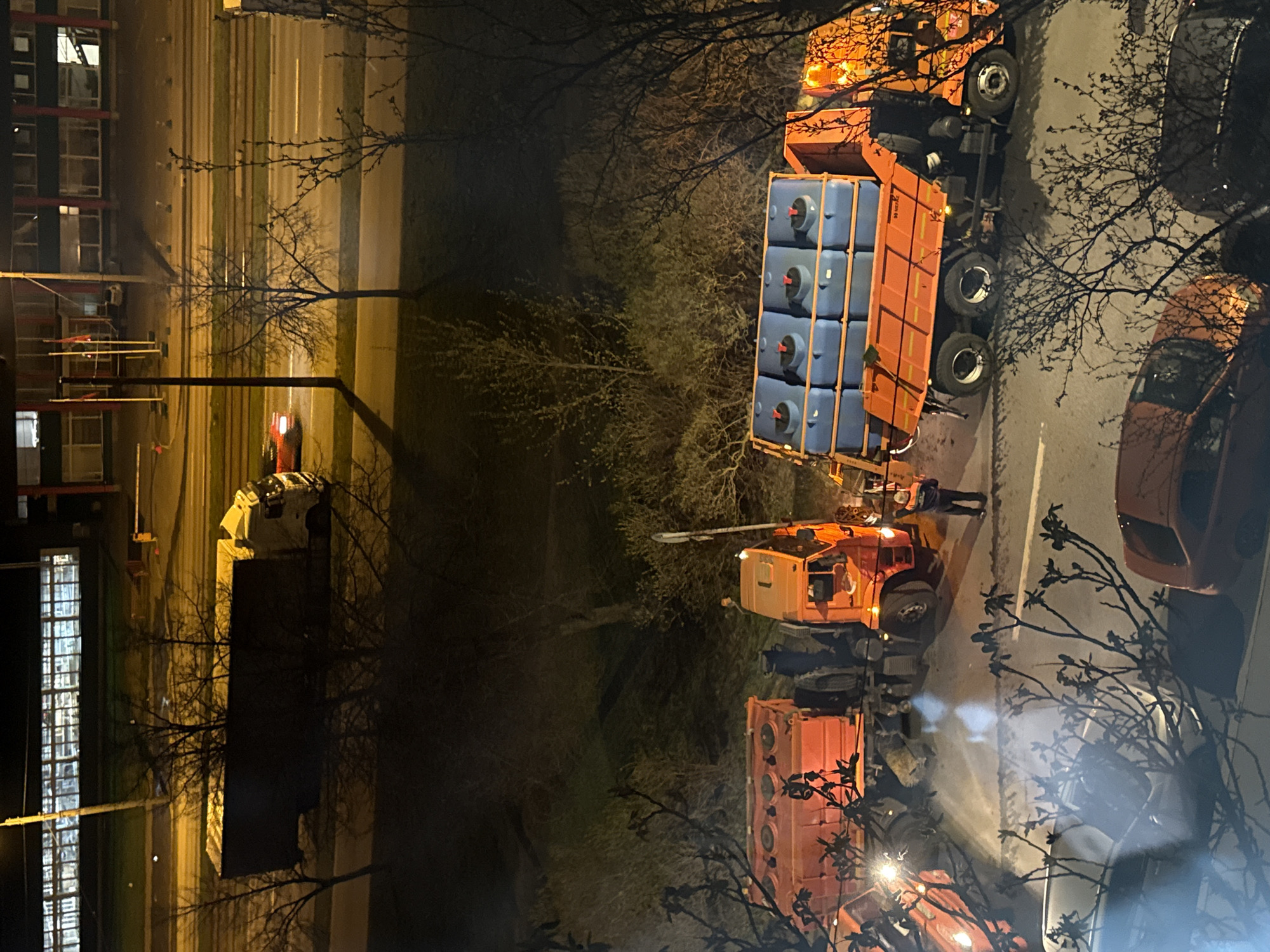 The image depicts a nighttime urban scene, likely in Saint Petersburg, Russia, illuminated by artificial light sources from vehicles and streetlights. The primary subjects are several large orange municipal vehicles parked on a street.
A prominent orange truck, possibly a flatbed, is loaded with multiple cuboid blue containers, each fitted with a red spigot or valve. The word "WHEEL" is visible on the truck's side. An articulated crane arm extends from one of the orange vehicles towards a person standing on the street. This individual, presumably a worker, appears to be actively involved in operating or guiding the crane, suggesting an ongoing task related to handling the blue containers.
Another large orange vehicle is parked behind the first, with parts of a third orange vehicle visible further down the road. On the right side of the street, an orange car is parked, partially obscured by bare tree branches. Across the street, a white car is also parked. The trees, lacking leaves, suggest the season is either winter or early spring.
To the left of the street, a multi-story building is visible, featuring illuminated windows and what appears to be scaffolding or exterior structures. The road itself is marked with lanes and runs parallel to what seem to be tram tracks. The overall ambiance is dim, characterized by the functional lighting required for nighttime work or maintenance activities.