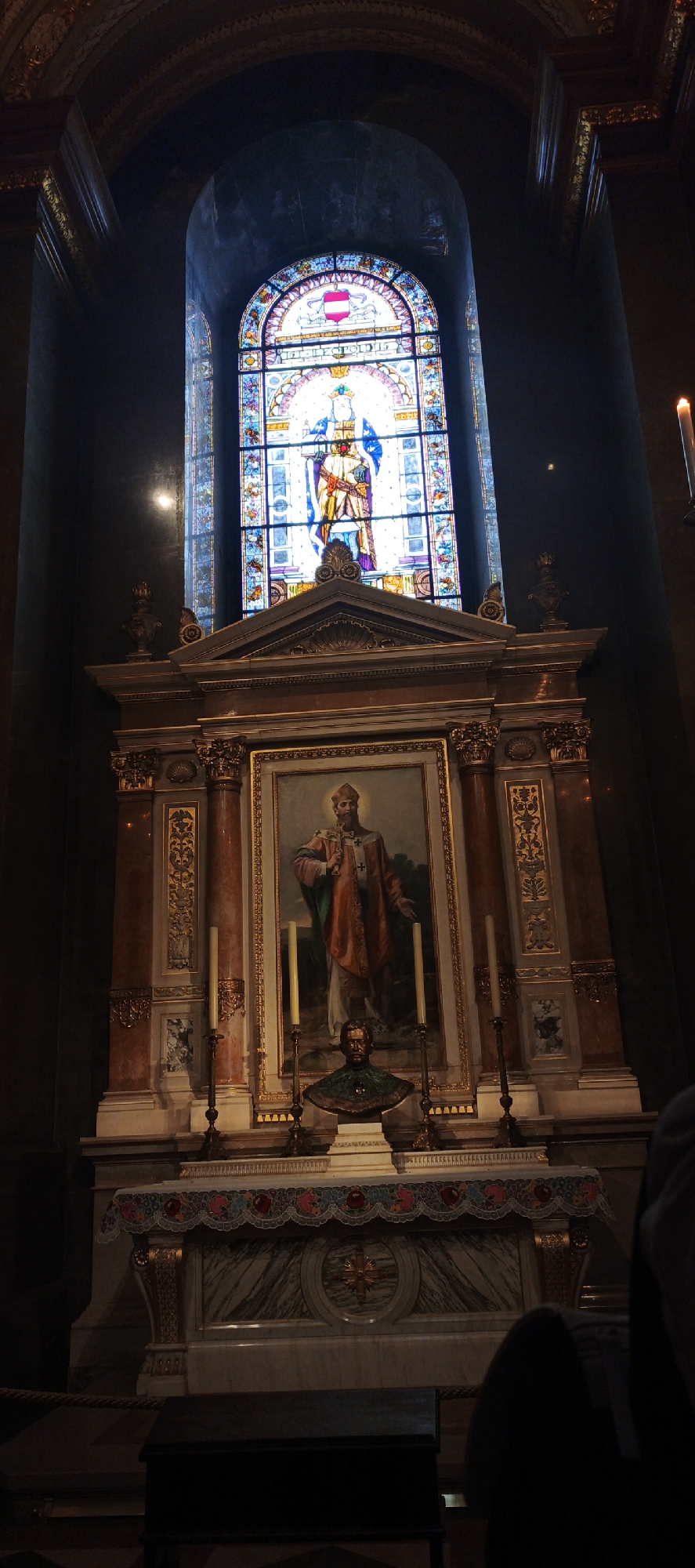 This image shows an altar in St. Stephen's Basilica in Budapest, Hungary. Above the altar is a stained-glass window depicting a king-like figure, likely Saint Stephen himself. The altar beneath displays a framed painting of a saintly figure with a beard and red robe. Below the painting is a bust, positioned on the altar. There are several tall, thin candles on the altar. The environment is dark, with some details obscured by the shadows.
