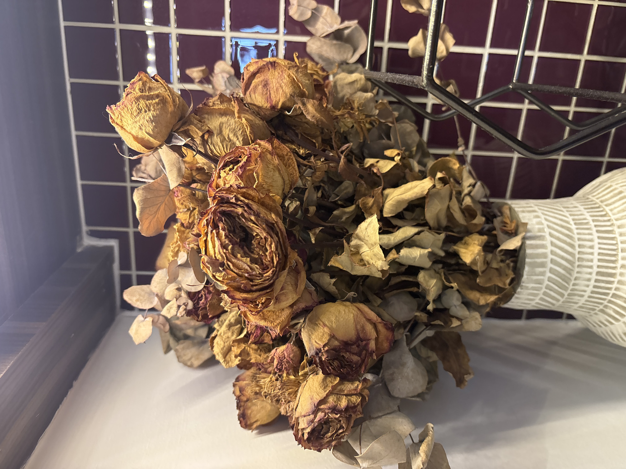 The image shows a close-up, eye-level, medium shot of a bouquet of dried roses and eucalyptus leaves in a decorative, white, textured vase. The flowers are predominantly orange and brown, with some faded pink hues on the petals. The background consists of a white wall on the left, and a tiled wall with a dark pink, almost burgundy color, on the right. Above the tiled wall, a dark wooden shelf is visible. A black wire rack is positioned in front of the tiled wall, partially obscuring the bouquet. The lighting appears to be artificial, casting shadows on the white wall and highlighting the textures of the flowers and vase. The scene is indoors and appears to be a still life arrangement. There are no people or animals present in the image. The image does not contain any information to suggest it is located in Dubai, United Arab Emirates.