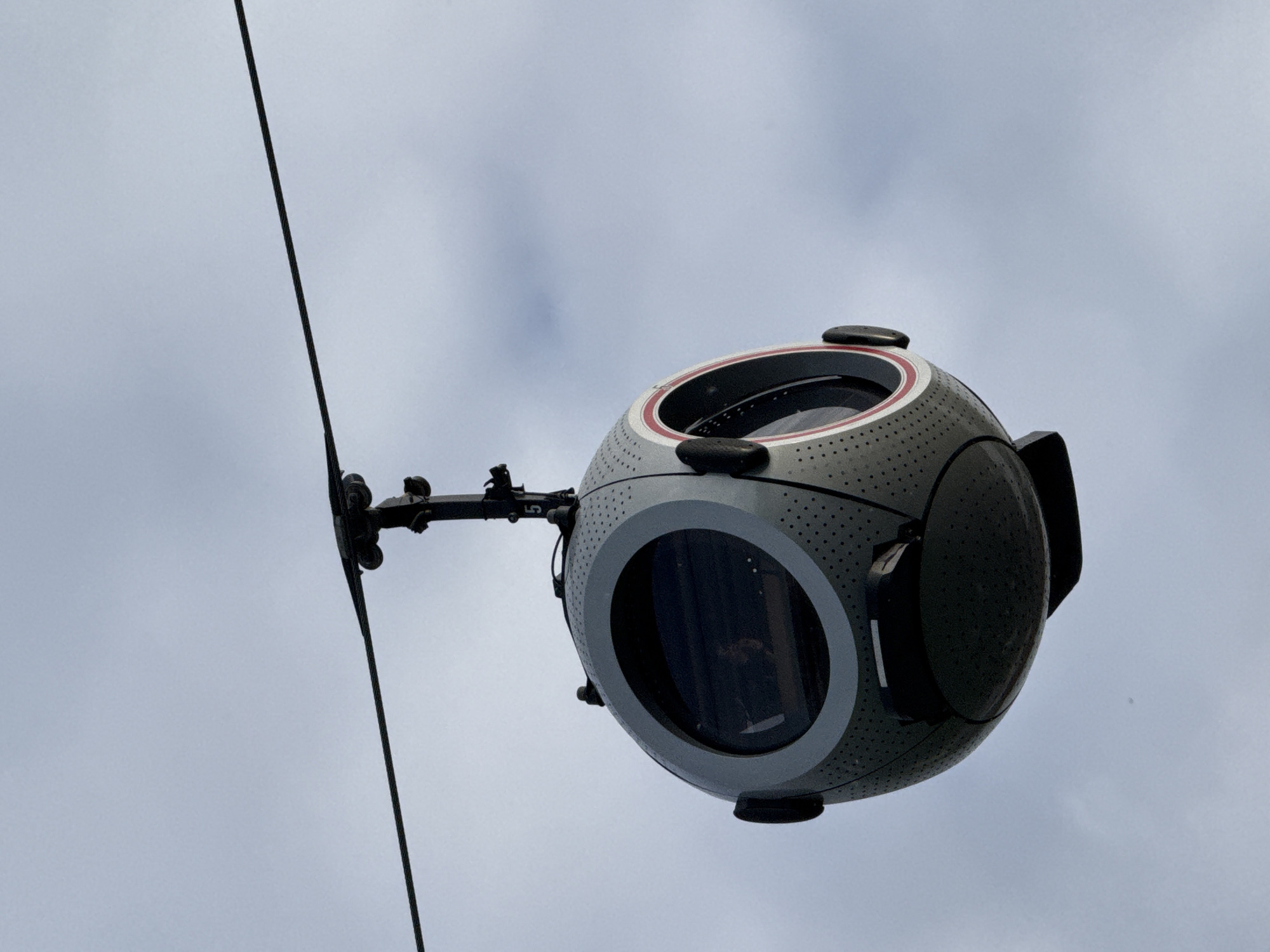 A futuristic, spherical gondola, primarily grey with black accents, is suspended in an overcast sky. It is attached to a thick, dark cable via a metallic, segmented arm. The cable runs diagonally from the upper left to the lower right of the frame, suggesting an aerial transport or monitoring system.
The gondola features a distinctive design with a perforated texture across its surface. It has several circular windows or lenses; one prominent circular window in the center is framed by a white and red ring. Other smaller, dark circular elements and protrusions are visible on its surface, resembling sensors or lights. The connecting arm, which holds the gondola to the cable with visible wheels or rollers, has the number "5" faintly visible on its side.
The sky is a uniform, light grey, indicating an overcast or cloudy day. The image captures the object from a low angle, looking upwards. The overall impression is of advanced technology, possibly for surveillance or passenger transport, set against a subdued sky. This scene takes place in Moscow, Russia.