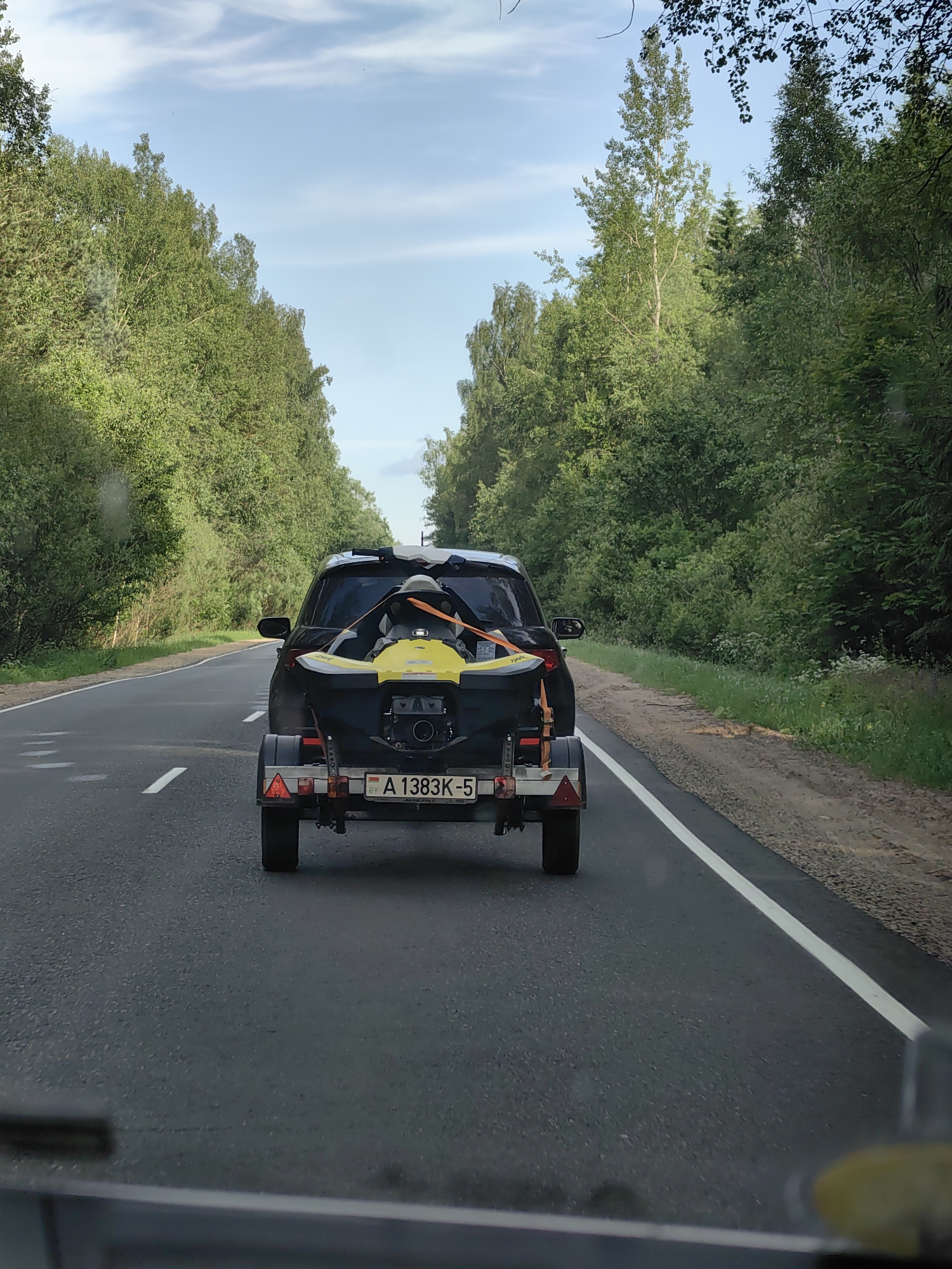 A black car with a trailer carrying a yellow jet ski drives on a paved road lined with trees. The license plate on the trailer reads "A 1383K-5". The sky is blue with some white clouds. The scene appears to be on a rural road, possibly in the vicinity of Хаценчыцкі сельскі Савет, Belarus.