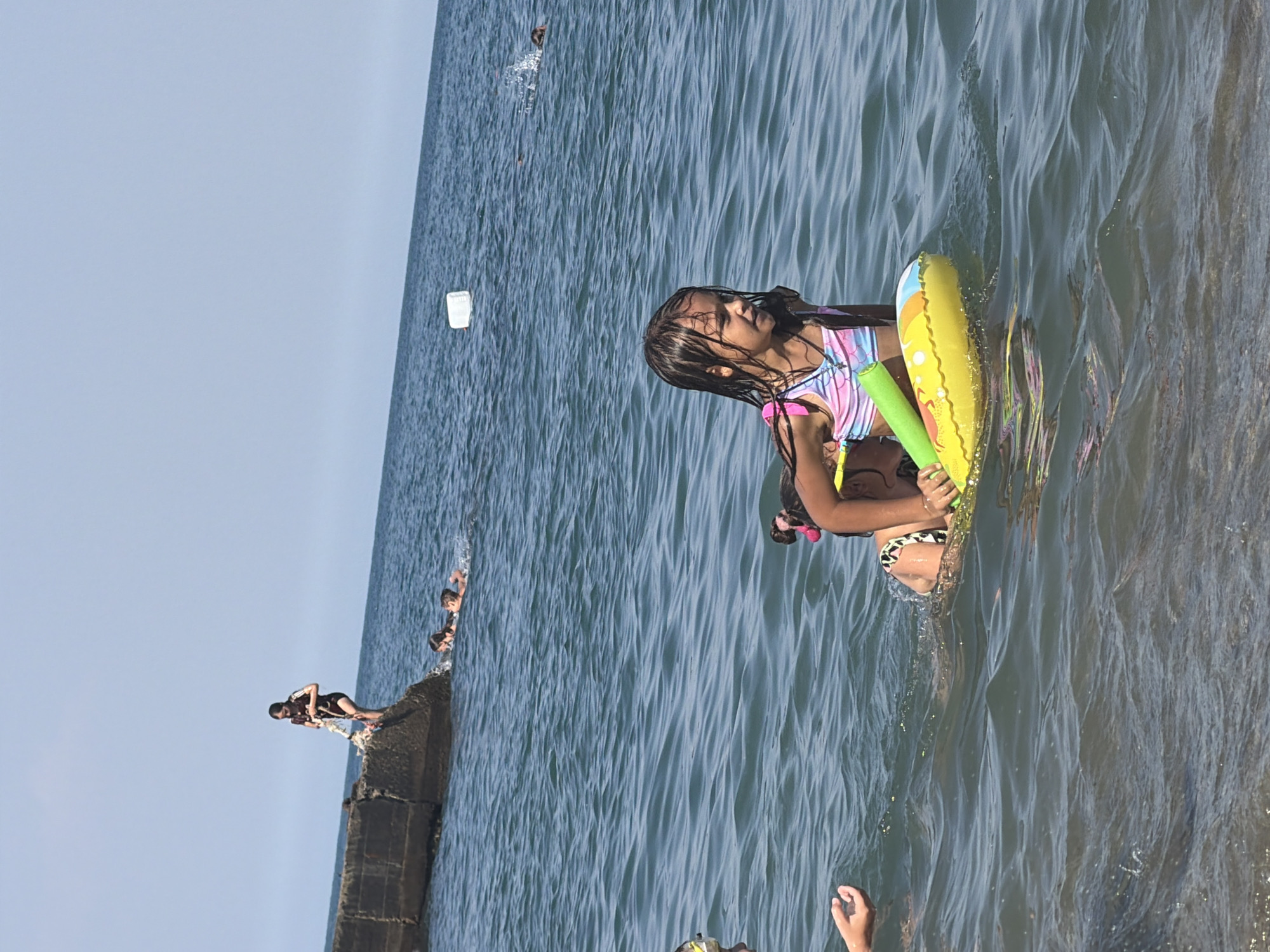 The image captures a bright, sunny day at a body of water, likely a sea, with people enjoying recreational activities. In the foreground, a young girl is partially submerged in the water, holding onto a yellow inflatable ring and a green pool noodle. Her wet hair and swimsuit suggest she has been swimming. Behind her, the water is a vibrant blue, with small waves rippling across the surface.
Further in the background, a concrete structure, possibly a pier or seawall, juts out into the water. Several people are gathered on this structure, and some are in the process of jumping into the water. One person in a dark swimsuit is poised to dive. Other individuals are visible in the water near the structure, seemingly enjoying the swim. The sky is a clear, pale blue, indicating fair weather. The overall atmosphere appears to be one of carefree summer fun. No visible text is present in the image.