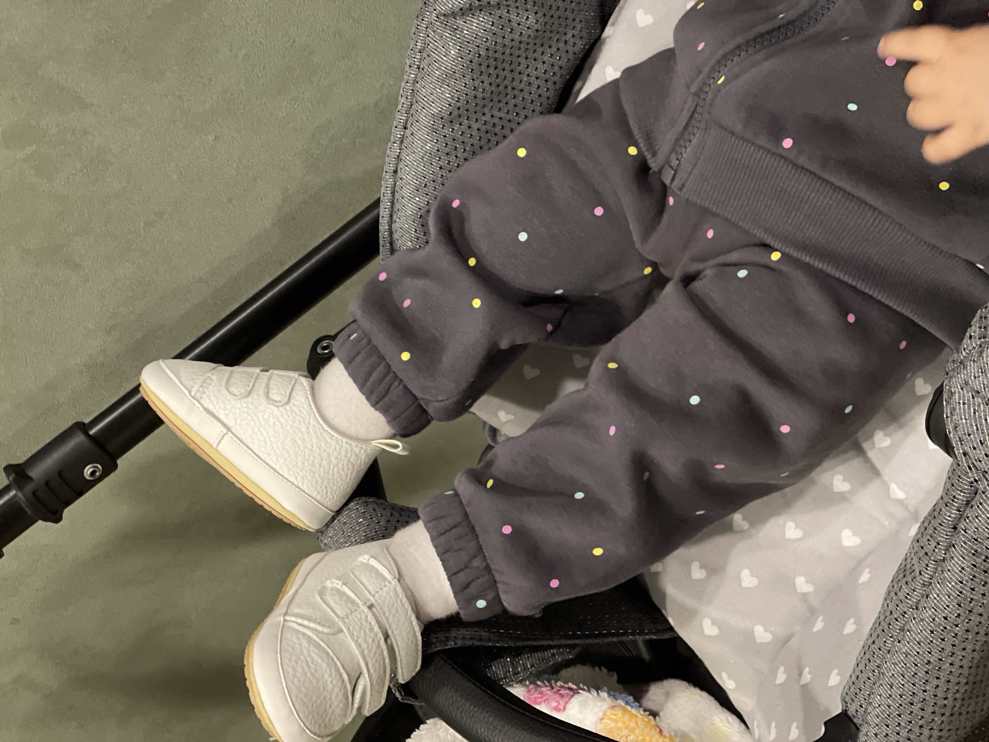 A close-up shot focuses on a baby's legs and feet, resting in a dark gray stroller. The baby is dressed in dark gray sweatpants patterned with colorful polka dots in yellow, pink, and light blue. They wear white socks and soft-soled white baby shoes, which have a textured appearance and yellowish soles. A blurred baby's hand is partially visible on the upper right side of the frame.
The stroller's interior is lined with a light gray fabric featuring a pattern of small white hearts. The stroller's exterior features a textured, dark gray material, possibly with a subtle sheen. A black structural bar or handlebar from the stroller is visible on the left side of the image. A plain, light green wall or surface serves as the background. A colorful, soft object, possibly a toy or blanket, peeks from the bottom right corner of the stroller. The indoor setting suggests the baby is resting comfortably in the stroller. No visible text is present.