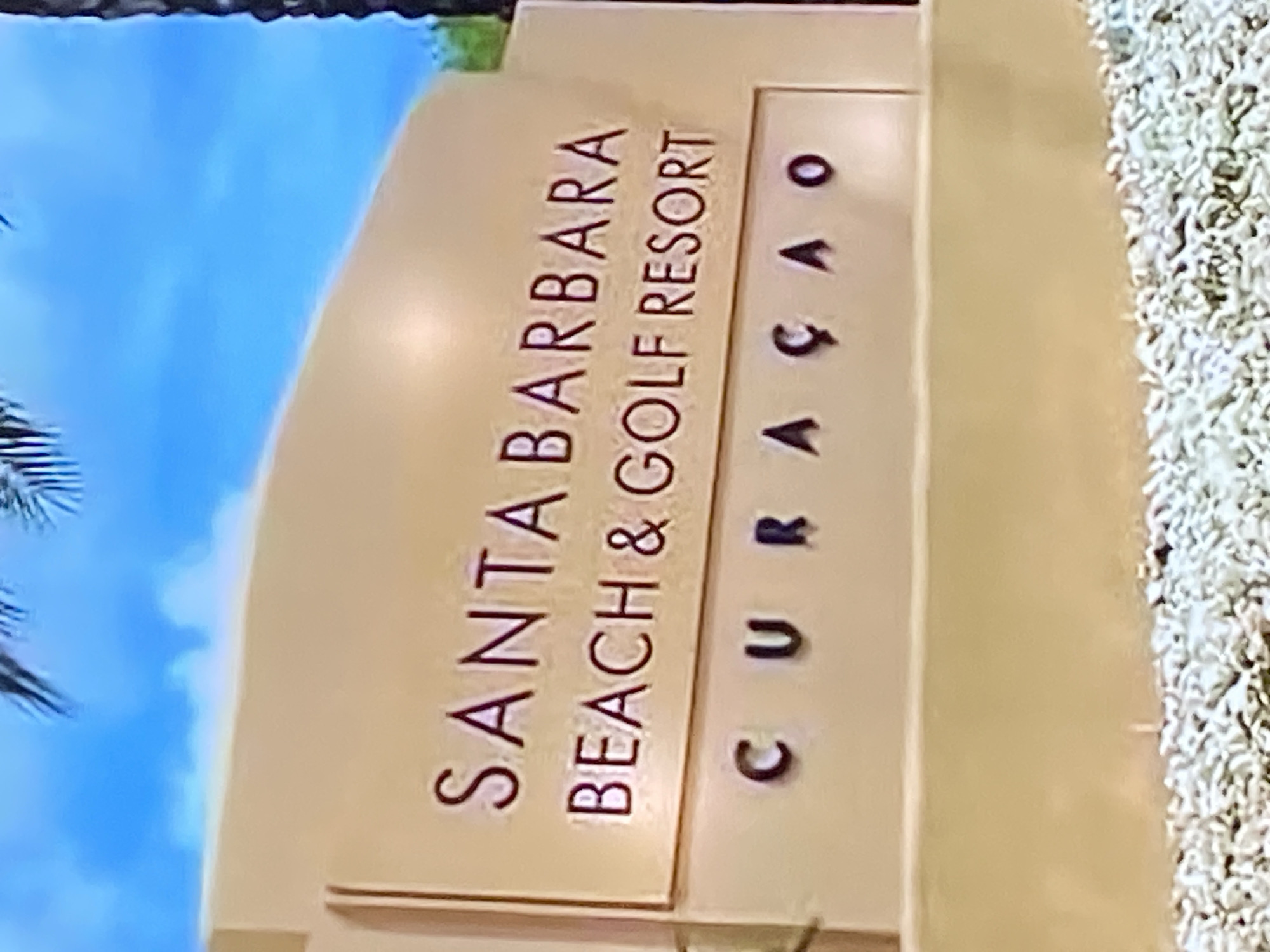 A light tan, three-tiered sign displays the words "SANTA BARBARA BEACH & GOLF RESORT CURAÇAO" in dark brown lettering. The top tier of the sign is partially obscured by a palm frond. The sign is set against a bright blue sky with some white clouds and a hint of green foliage on the right side. The bottom of the sign rests on a bed of small white and grey gravel.