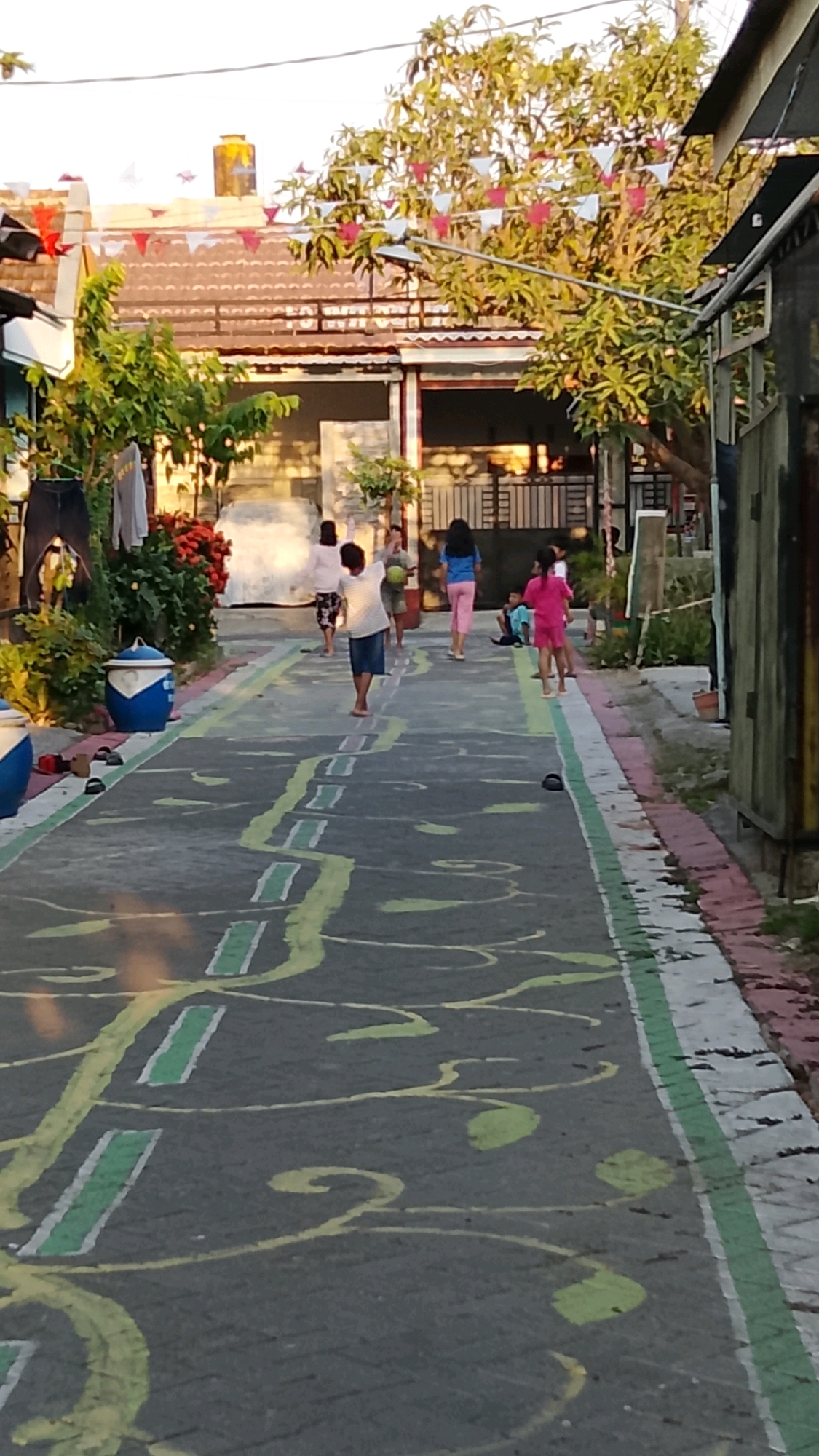 In Banjarsari, Indonesia, children are playing a game on a street that has been painted with a hopscotch-like design. The game appears to be hopscotch or a similar jumping game, with colored squares and lines painted on the asphalt. Several children, mostly young, are scattered across the street, some mid-jump, others observing. The environment is a narrow street with houses lining both sides. Laundry hangs from lines outside one of the houses, and potted plants are visible. Trees with red and white triangular flags strung between them suggest a festive occasion or decoration. The sky is clear, indicating daylight. The overall scene depicts a common urban setting where children utilize public spaces for recreation.