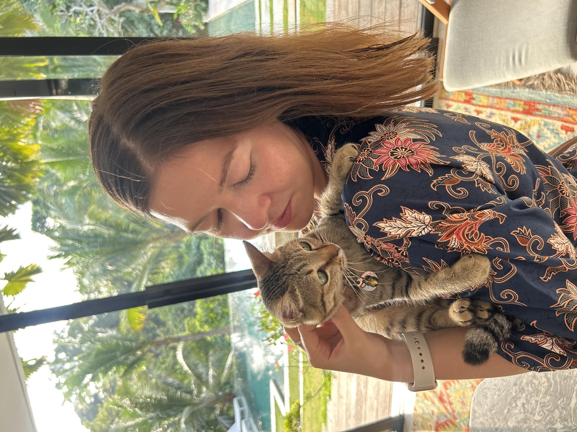 A medium shot captures a young woman with light brown hair holding a tabby cat in her arms. The woman, with her eyes closed, is gently stroking the cat. The cat, with green eyes, looks forward with its ears perked. The background features a swimming pool and lush greenery, suggesting an outdoor setting. The woman wears a dark blue dress with a floral pattern. The scene appears to be indoors with large windows offering a view of the outdoors, possibly in a tropical location. No specific location in Kediri, Indonesia, can be determined from the image.