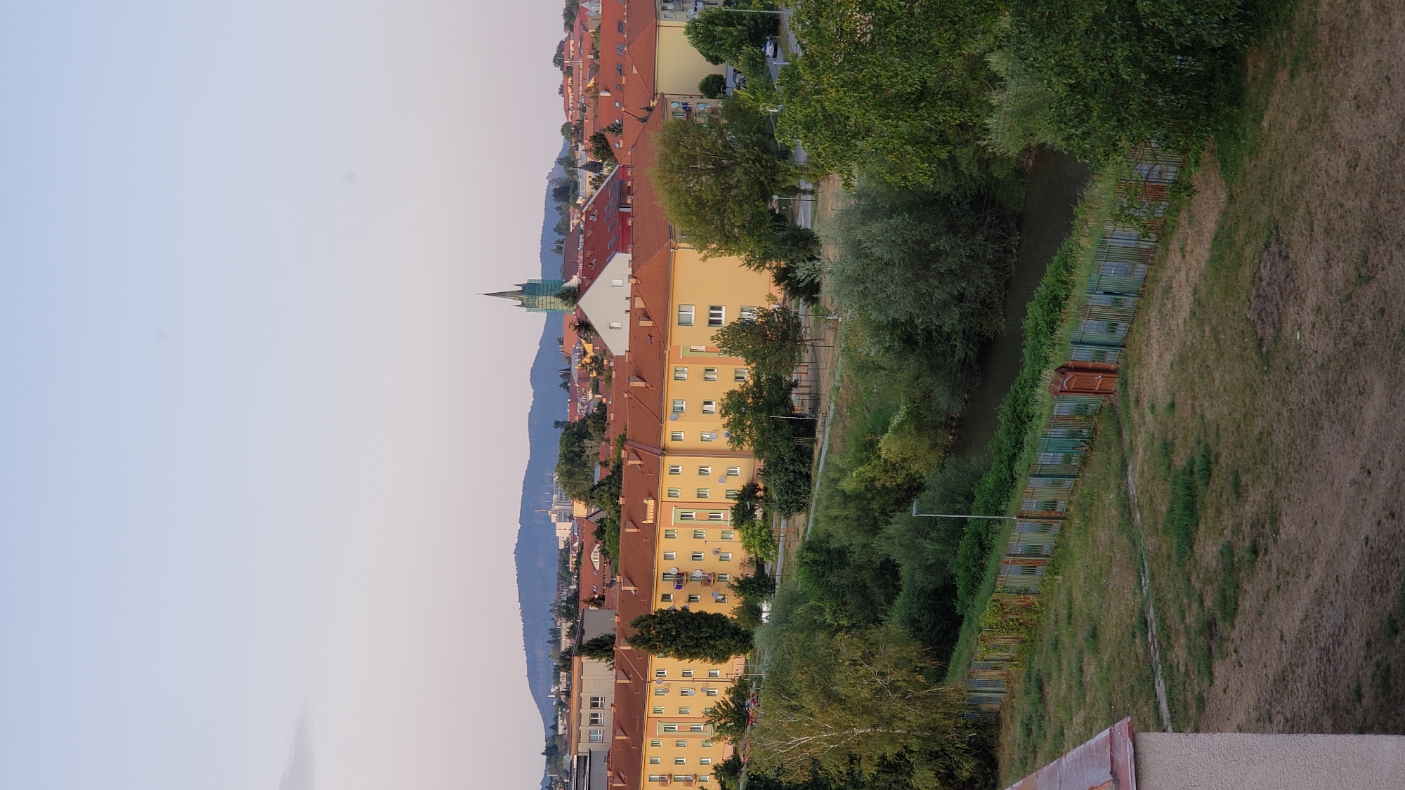 The image depicts a panoramic view of a cityscape with a residential area in the foreground and a distant hill. In the middle ground, a long, yellow apartment building with orange-tiled roofs stretches across the frame. Trees of various sizes are interspersed throughout the scene, with lush green foliage. A small river or canal is visible in the lower part of the image, bordered by a green fence and a grassy embankment. In the background, more buildings with red roofs are scattered, and a distinctive green church steeple rises above the rooftops. A dark, tree-covered hill with a flat top dominates the horizon. The sky is a pale blue, indicating it is likely late afternoon or early evening. The foreground shows a section of a building with a reddish-brown roof and a textured light-colored wall. The overall impression is of a calm, urban environment with a touch of nature. The image appears to be taken from an elevated position, possibly a balcony or rooftop.