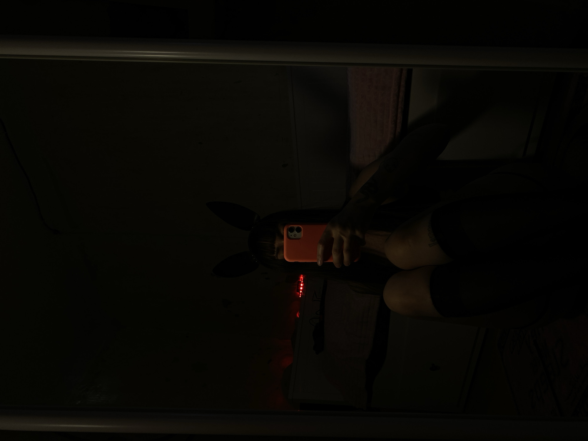 A reflection is captured in a mirror, revealing a person wearing bunny ears and holding an orange smartphone in front of their face. Red string lights and a white notepad or book are also visible in the reflection. The background is dark, obscuring details of the room and its location. The location in Votkinsk, Russia, cannot be determined from the image.