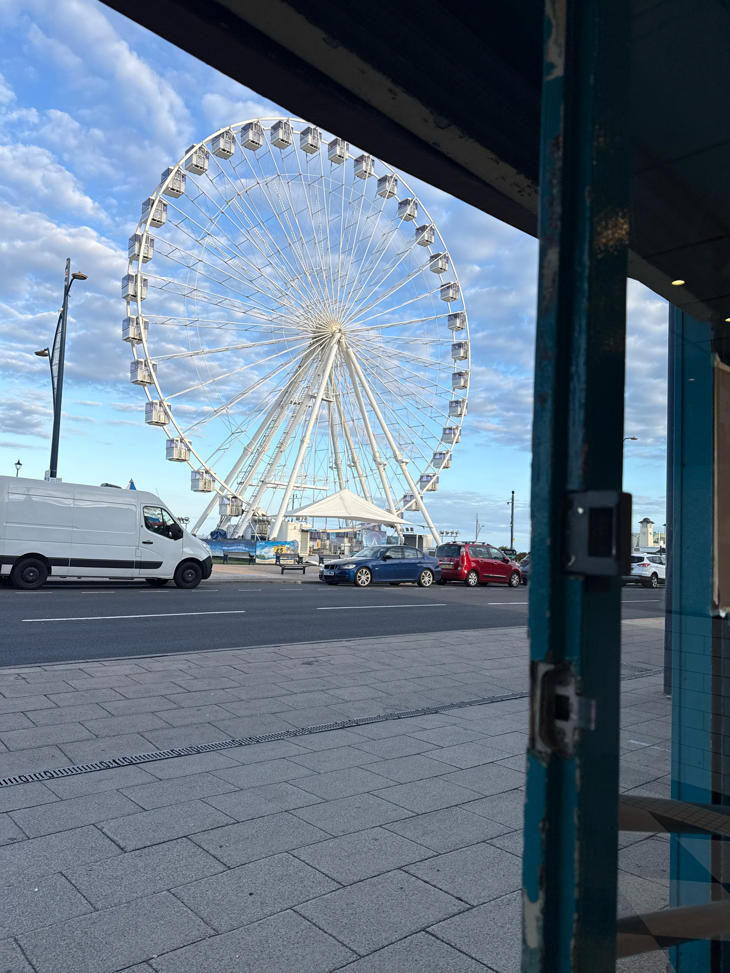 The image depicts a Ferris wheel in Great Yarmouth, United Kingdom, viewed from behind a partially visible blue metal structure. The Ferris wheel, a prominent attraction, is large and white with numerous gondolas. In the foreground, a white van is parked on the left side of a road, with a blue car and a red car further down the road. The ground is paved with grey tiles, and a metal drain runs across it. The sky is blue with scattered white clouds, and a street lamp is visible on the left. The overall scene suggests an outdoor location, possibly a promenade or public area near a waterfront, given the presence of the Ferris wheel and the general seaside atmosphere associated with Great Yarmouth. The blue structure in the foreground appears to be part of a building's facade or a kiosk, framing the view of the Ferris wheel. There are no people clearly visible engaging in any specific actions.