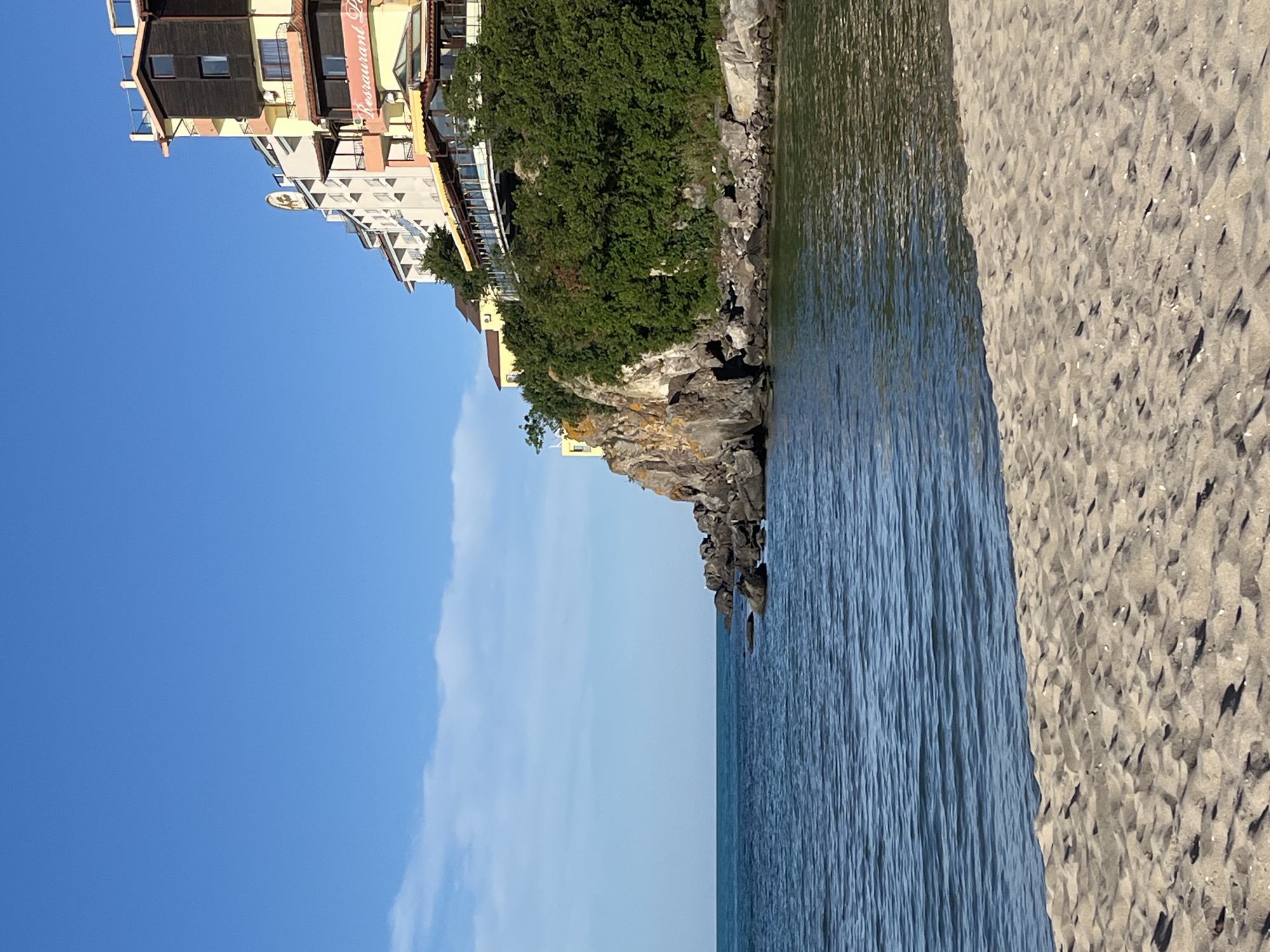 A vertical, eye-level shot captures a serene beach scene in Sozopol, Bulgaria, under a clear blue sky. In the foreground, a sandy beach transitions into the calm, dark blue waters of the sea. The water is rippled, reflecting the sunlight.
To the right, a rocky cliff rises, densely covered with lush green trees. Perched atop the cliff is a multi-story hotel or apartment complex with various architectural styles and colors, including yellow, white, and dark wood elements. Some balconies overlook the sea, and a sign with "Restaurant P" is visible in red lettering on one of the yellow sections. The buildings appear to be built into the cliff face, offering a dramatic juxtaposition of nature and architecture. The overall impression is one of a tranquil coastal resort.