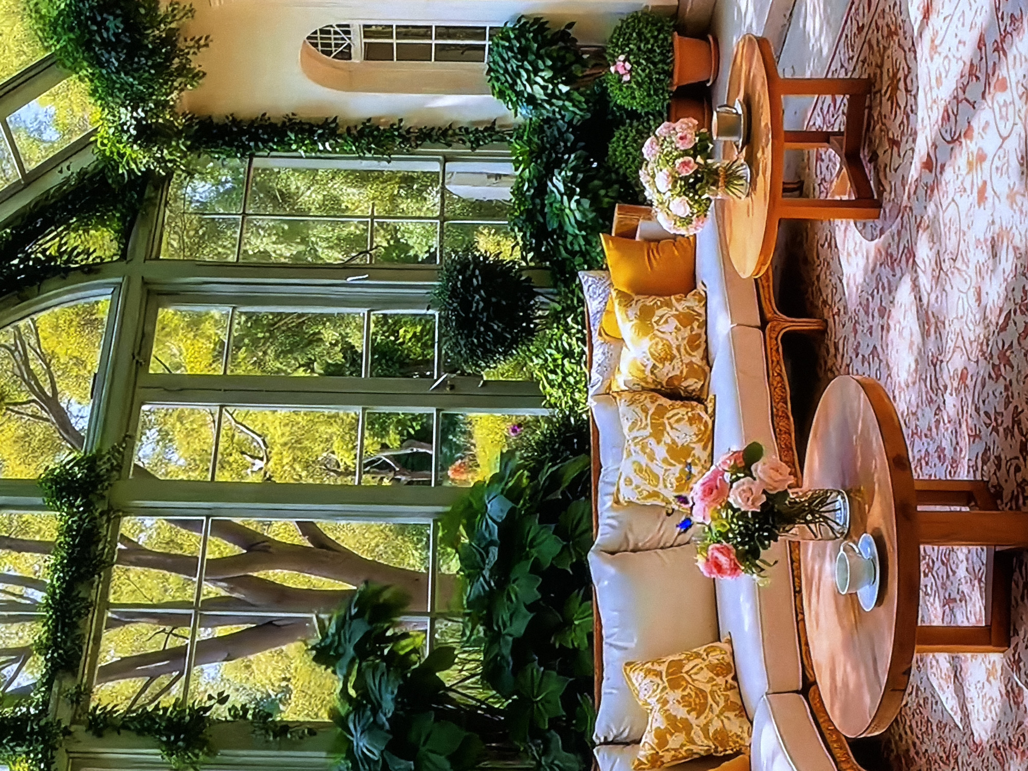 This media file captures a serene and elegantly furnished sunroom or conservatory located in Sofiivska Borshchahivka, Ukraine. The dominant feature is a wall of large, multi-paned windows, framed by light green sashes and adorned with lush green vines, offering an expansive view of a vibrant outdoor landscape filled with green trees and dense foliage.
Inside the sunroom, the setting features comfortable light-colored sofas or benches, generously appointed with decorative cushions in yellow and white patterns, alongside solid yellow throw pillows. Two round, light-wood side tables are placed adjacent to the seating. Each table holds a clear glass vase brimming with fresh pink roses and other flowers. A white teacup and saucer are also visible on one of the tables. Various potted green plants, including neatly trimmed topiaries and larger leafy plants, are strategically arranged throughout the space, enhancing the indoor-outdoor ambiance. High on the back wall, an arched window with white grilles allows additional light.
The scene is brightly lit by natural sunlight, suggesting daytime, possibly morning or afternoon, with sunbeams creating dappled patterns across a light-colored, patterned rug that covers the floor. The overall atmosphere is one of tranquility, comfort, and a close connection to nature. No people are present in the image.