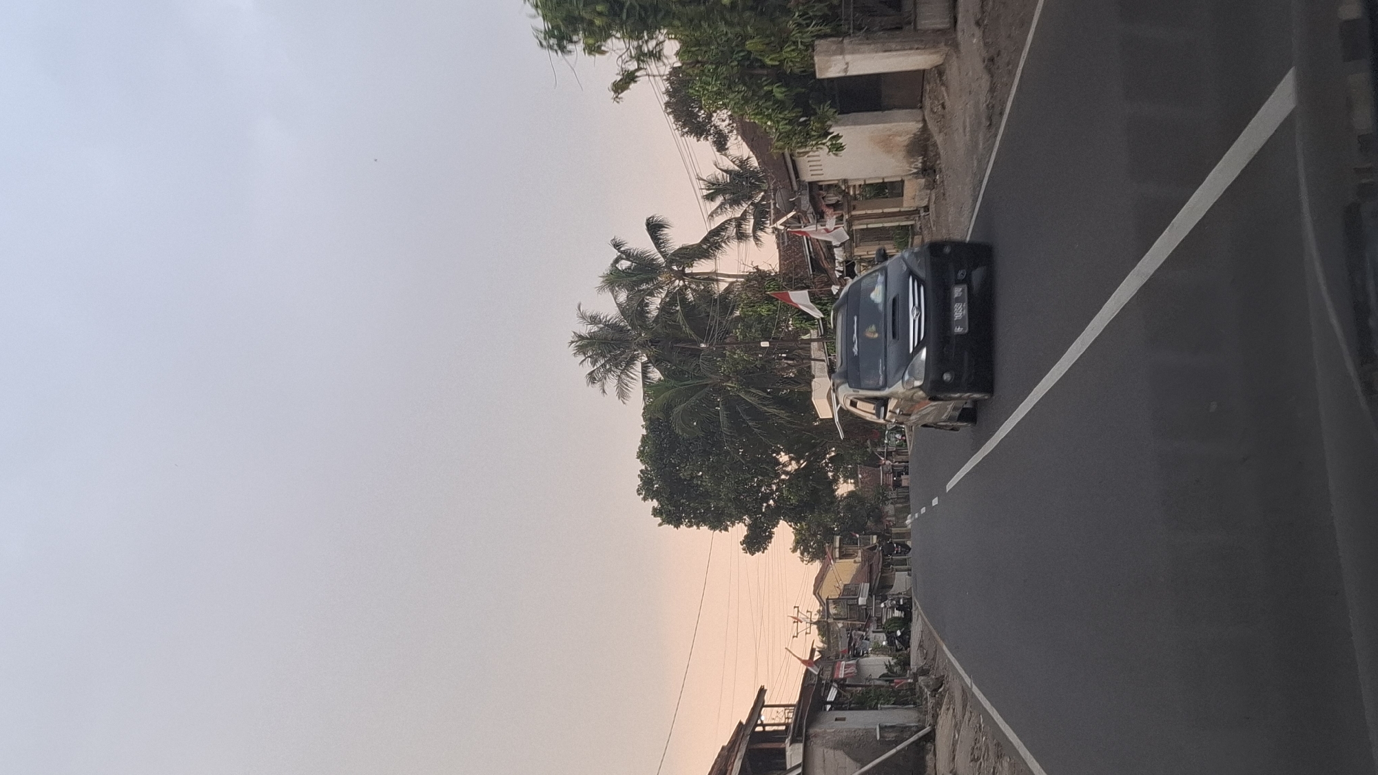 The image depicts a street scene in Susukan, Indonesia, during the late afternoon or early evening, as indicated by the soft, warm light of the setting sun. A black minivan is driving on the asphalt road, which is marked with a white dashed line in the center and a solid white line on the right. The minivan has its headlights on and a license plate that is partially visible. On the left side of the road, there are several buildings, some appearing to be houses with white walls and tiled roofs. There are also some motorcycles parked along the roadside. Trees, including several palm trees, line the street and are visible behind the buildings. Two Indonesian flags are displayed on poles in front of some of the houses, suggesting a national celebration or commemoration. The sky is a pale, hazy blue with hints of pink and orange near the horizon, typical of a sunset. Power lines are strung across the upper portion of the image, supported by utility poles. The overall impression is that of a quiet, residential street in a tropical or subtropical region.