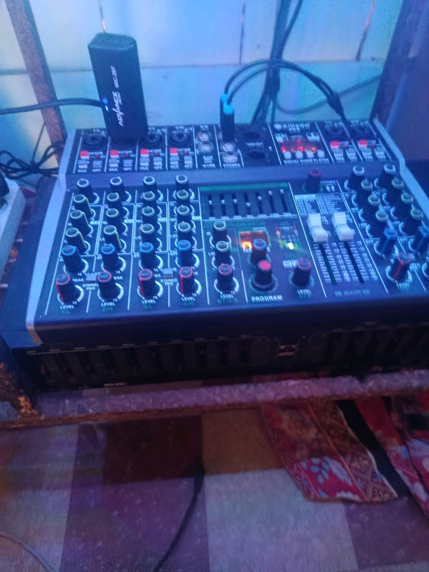 An audio mixer with various knobs and sliders is placed on a wooden surface. A black device labeled "ADVANCE MIC-207" is plugged into the mixer. The mixer has multiple input channels, each with gain, EQ, and level controls. Some indicator lights are visible on the mixer. The surrounding environment appears to be dimly lit, with blue and purple hues suggesting artificial lighting. The surface beneath the mixer is covered with a patterned cloth. The scene is indoors, possibly a studio or performance space. The specific location in Trisobo, Indonesia, is not discernible from the image.