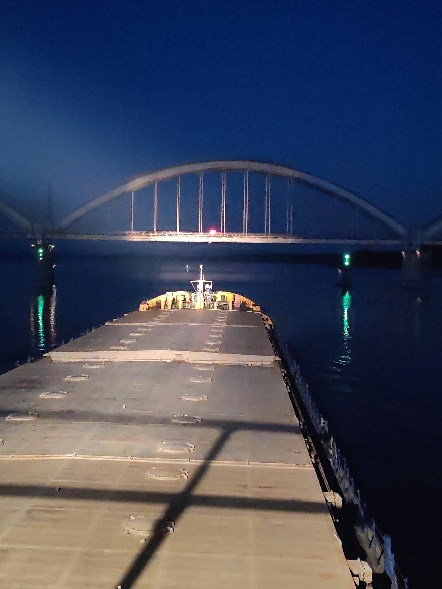 This forensic image captures a nighttime scene from a vessel navigating a waterway in Gladyshevo, Russia. The perspective is from an elevated position on the ship, looking forward over its expansive, flat deck towards an arched bridge.
The primary subject in the foreground is the foredeck of a large cargo ship, likely a bulk carrier, characterized by numerous rectangular hatch covers. A distinct shadow, possibly from an overhead structure or railing, extends diagonally across the deck from the upper left. A low-profile structure is visible at the forwardmost point of the deck.
In the mid-ground, a prominent white or light-colored tied-arch bridge spans the waterway. Vertical suspenders connect the arch to the bridge's deck. Red navigation lights are visible on the bridge structure. Beneath the bridge, on the dark, reflective water, two distinct green navigation lights are visible on what appear to be buoys or pylons, casting bright green reflections onto the calm surface.
The surrounding environment consists of dark blue twilight sky, indicating night. The water is dark and still, reflecting the various light sources. No individuals are visible in the scene.