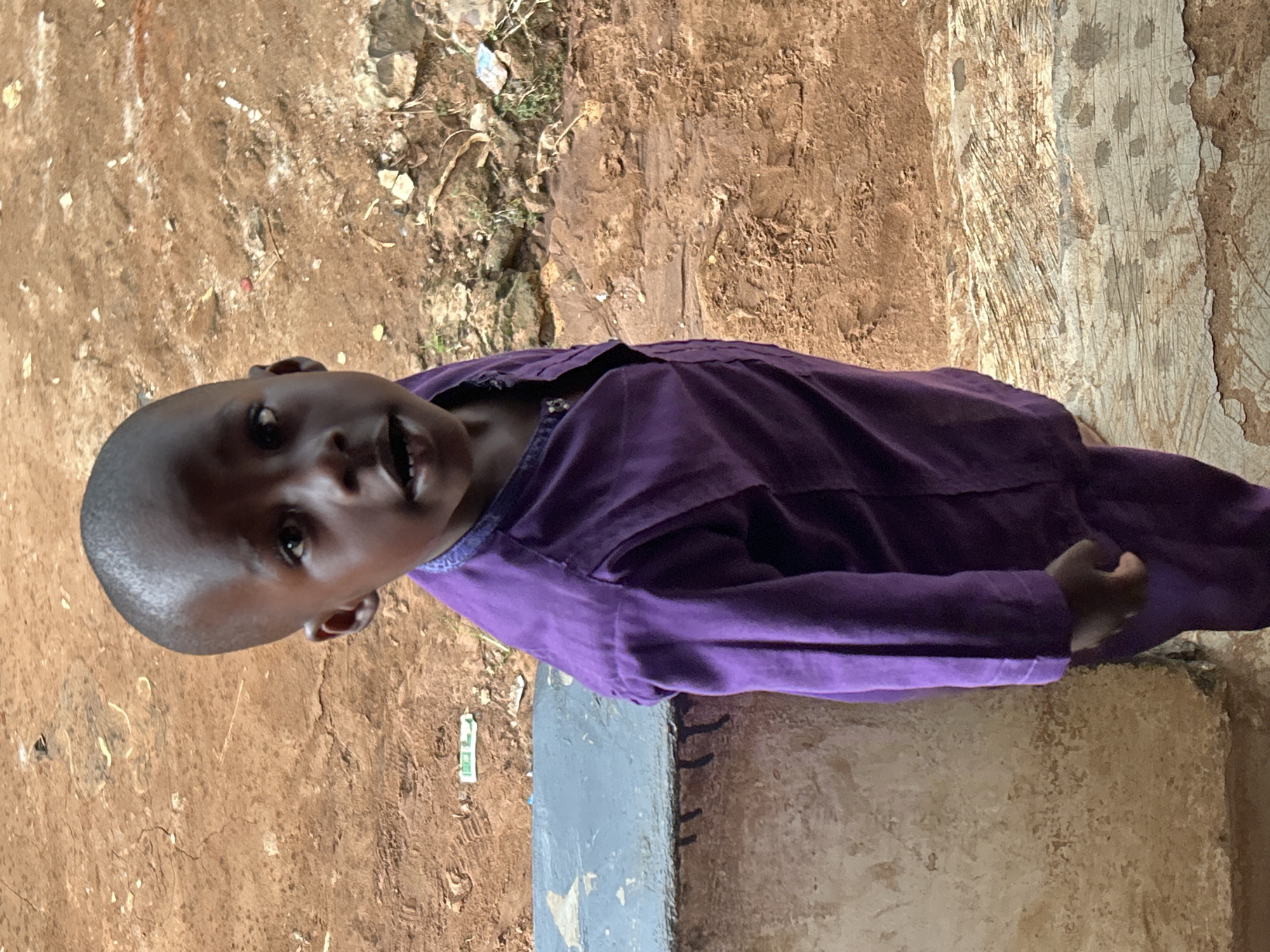 A young child with dark skin and a shaved head is wearing a purple shirt and standing outdoors. The child's gaze is directed towards the camera. The ground appears to be a mixture of bare earth and concrete. The city and country are unknown.