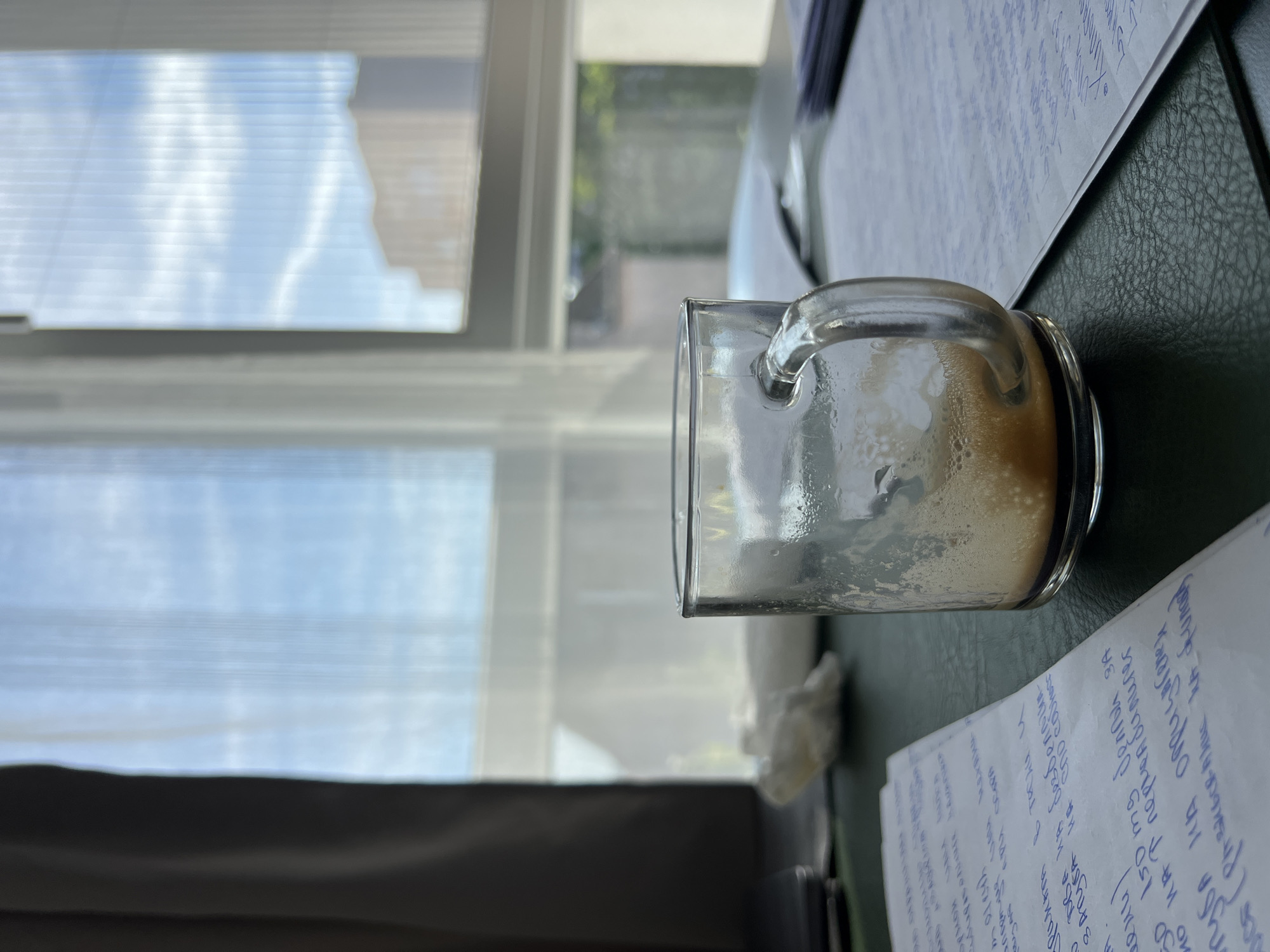 The image shows a desk with a partially empty glass mug of coffee. The mug is made of clear glass with a handle and contains a frothy brown liquid at the bottom. In the background, a window with white translucent curtains reveals a bright sky with clouds and a building. There are papers with handwritten notes on the desk, some with blue ink. A crumpled piece of white tissue is also visible on the desk. The lighting suggests it is daytime. The scene appears to be an indoor workspace, possibly an office or study. The image does not provide any specific visual cues to determine the exact location in Burgas, Bulgaria.