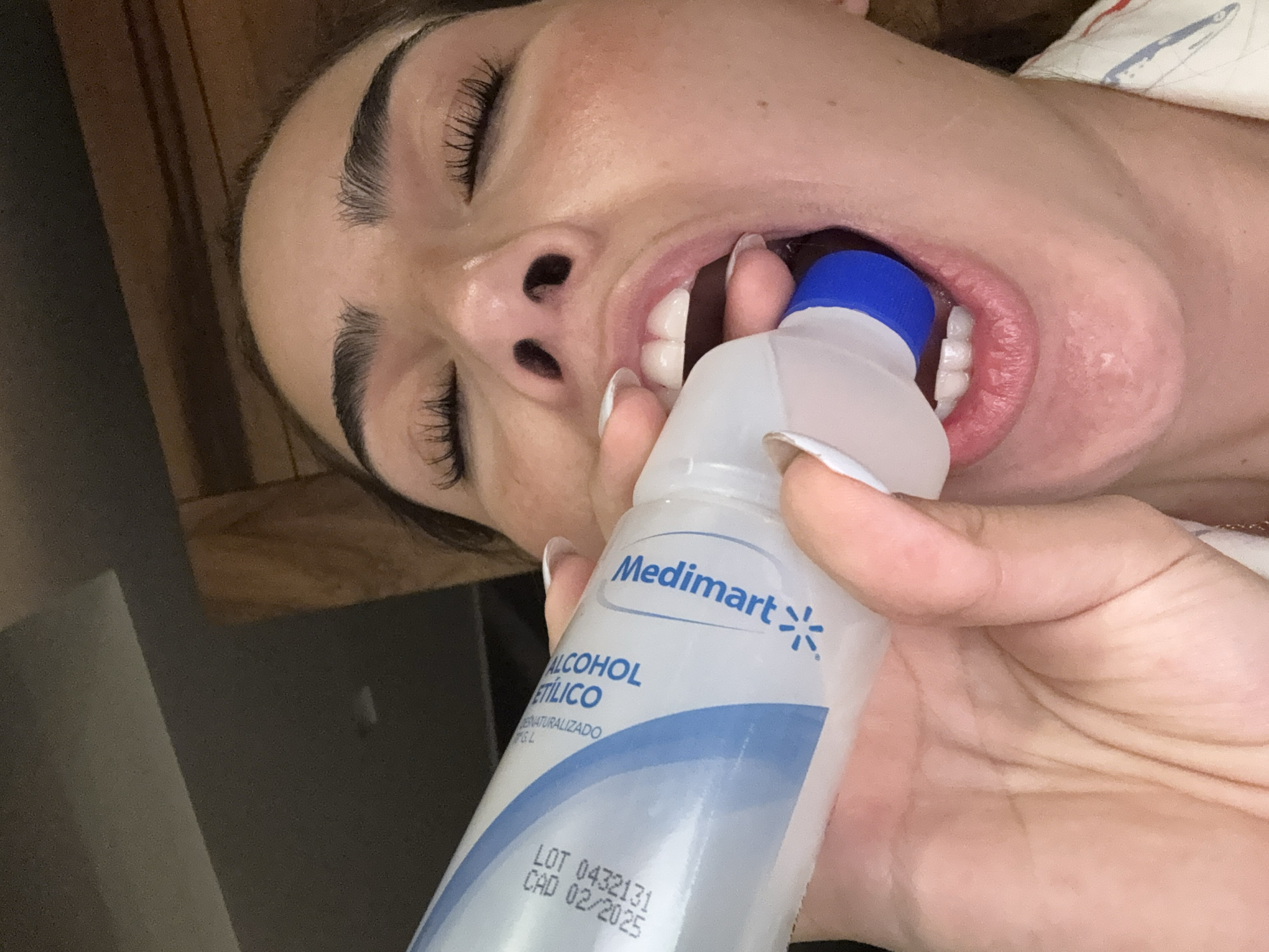 A woman with closed eyes and manicured fingernails is holding a bottle of "Medimart ALCOHOL ETILICO" up to her open mouth. The bottle has a blue cap and a lot number "LOT 0432131" with a CAD of "02/2025". The location in the city of San Bernardino Tlaxcalancingo, Mexico is not discernible from the image.