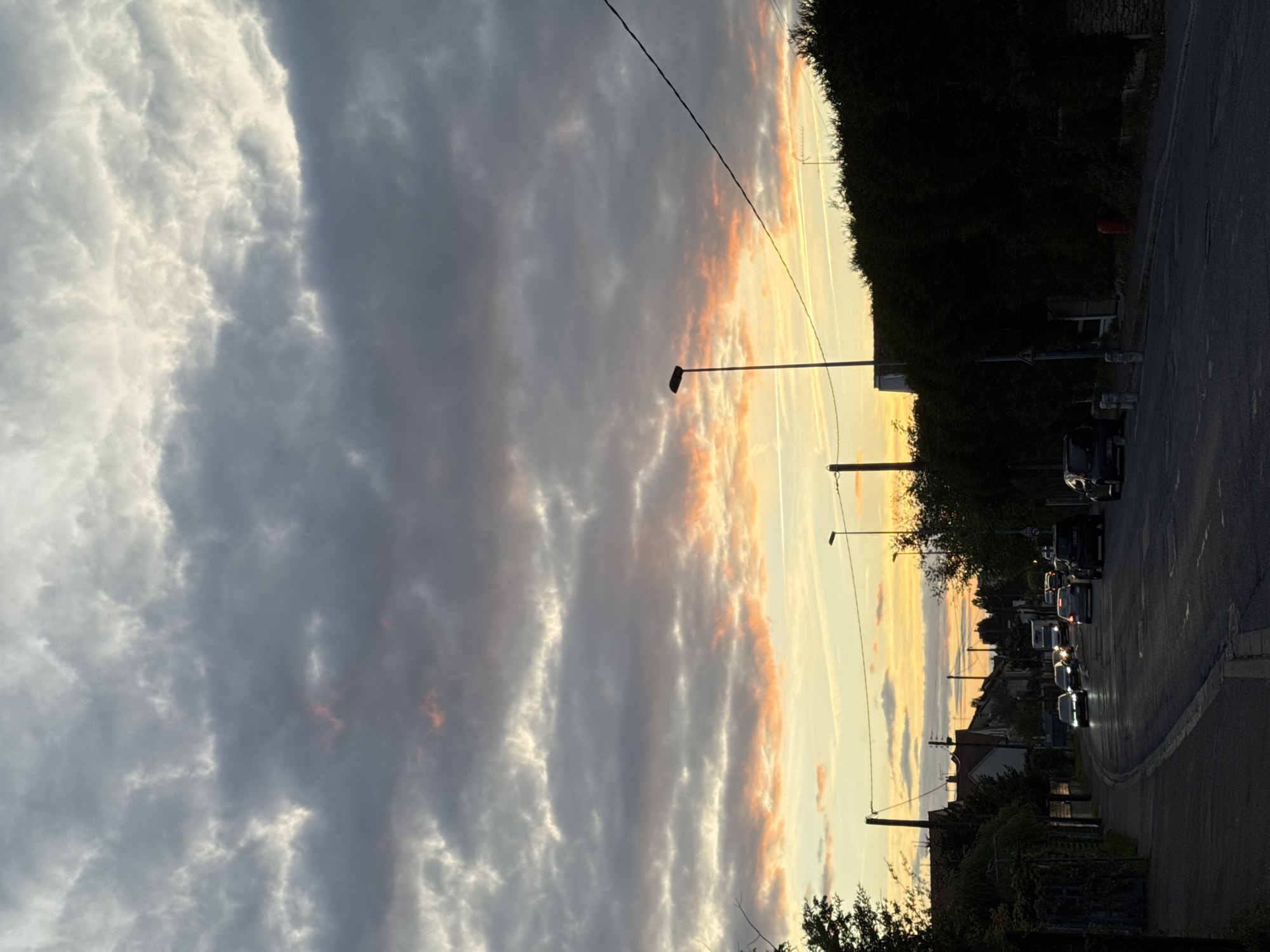 The image shows a street scene in Orsay, France, at dusk. The sky is filled with a dramatic cloudscape featuring hues of gray, white, and orange, suggesting a sunset. A street runs through the scene, lined with streetlights and power lines. On the right side of the road, there is dense foliage and parked cars. Further down the street, car headlights are visible, indicating traffic. Houses are visible in the distance. The image has a subdued, warm lighting, likely due to the setting sun.