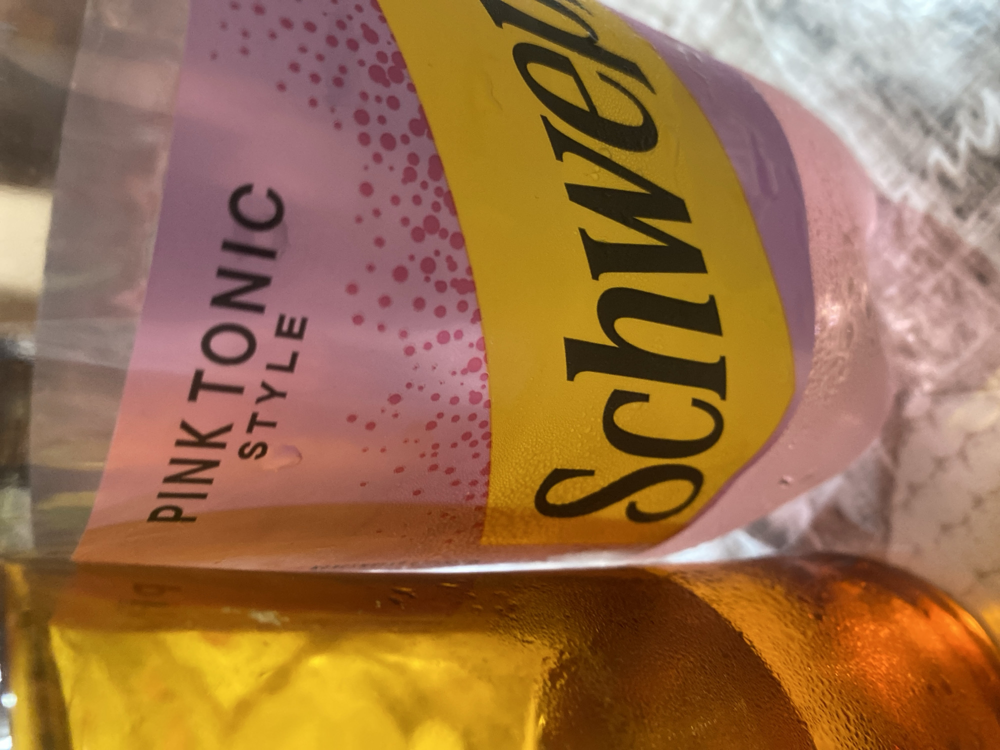 Two beverage containers are positioned side-by-side. To the left is a glass filled with a golden-amber liquid and ice. The glass has condensation on its exterior. To the right is a clear plastic bottle containing a pink liquid. The bottle has a label with pink and yellow sections. The pink section features text that reads "PINK TONIC STYLE" and is adorned with scattered pink dots. Below this, a prominent yellow band displays the black text "Schwepp". Condensation is visible on the exterior of the plastic bottle as well. The background appears to be a textured surface, possibly a tablecloth. The image does not provide any information about the city of Knezha, Bulgaria, or any individuals.
