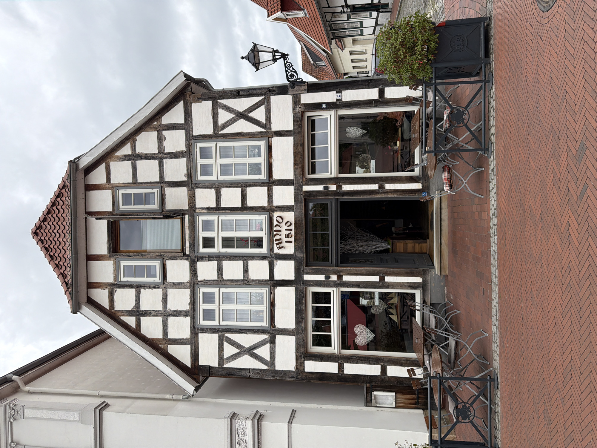 A wide, eye-level shot shows the facade of a historic, half-timbered building with an outdoor seating area in front. The building, with white infill and dark timber framing, has a prominent gabled roof with red tiles. A sign above the entrance reads "ANNO 1510." The entrance is a dark doorway with flanking windows. The windows on the upper floors are multi-paned and appear to be original. The ground floor features large display windows, one of which has a white heart-shaped decoration hanging inside.
In front of the building, on a red brick paved area, are several sets of outdoor tables and chairs. A black metal railing encloses a portion of the seating area. A large, dark planter with a green shrub sits to the right of the seating area. To the left of the building is a portion of an adjacent, lighter-colored building. A decorative black lamppost is mounted on the side of the half-timbered building, to the right of the entrance. The sky is overcast and gray. There are no people clearly visible in the scene.